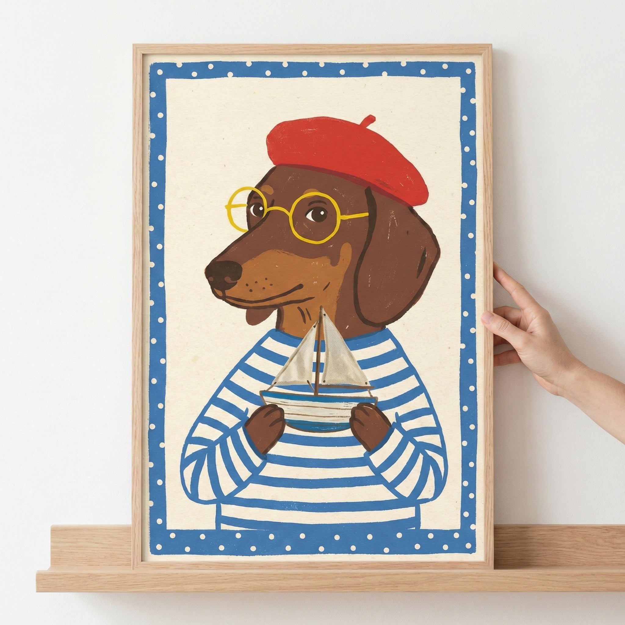 An illustration of a brown dachshund dog wearing a red beret, yellow round-framed glasses, and a blue and white horizontally striped shirt. The dog is holding a small toy sailboat in its paws. The artwork is set within a blue border featuring white polka dots and is displayed in a light-colored wooden frame sitting on a wooden shelf, with a person's hand visible on the right side.