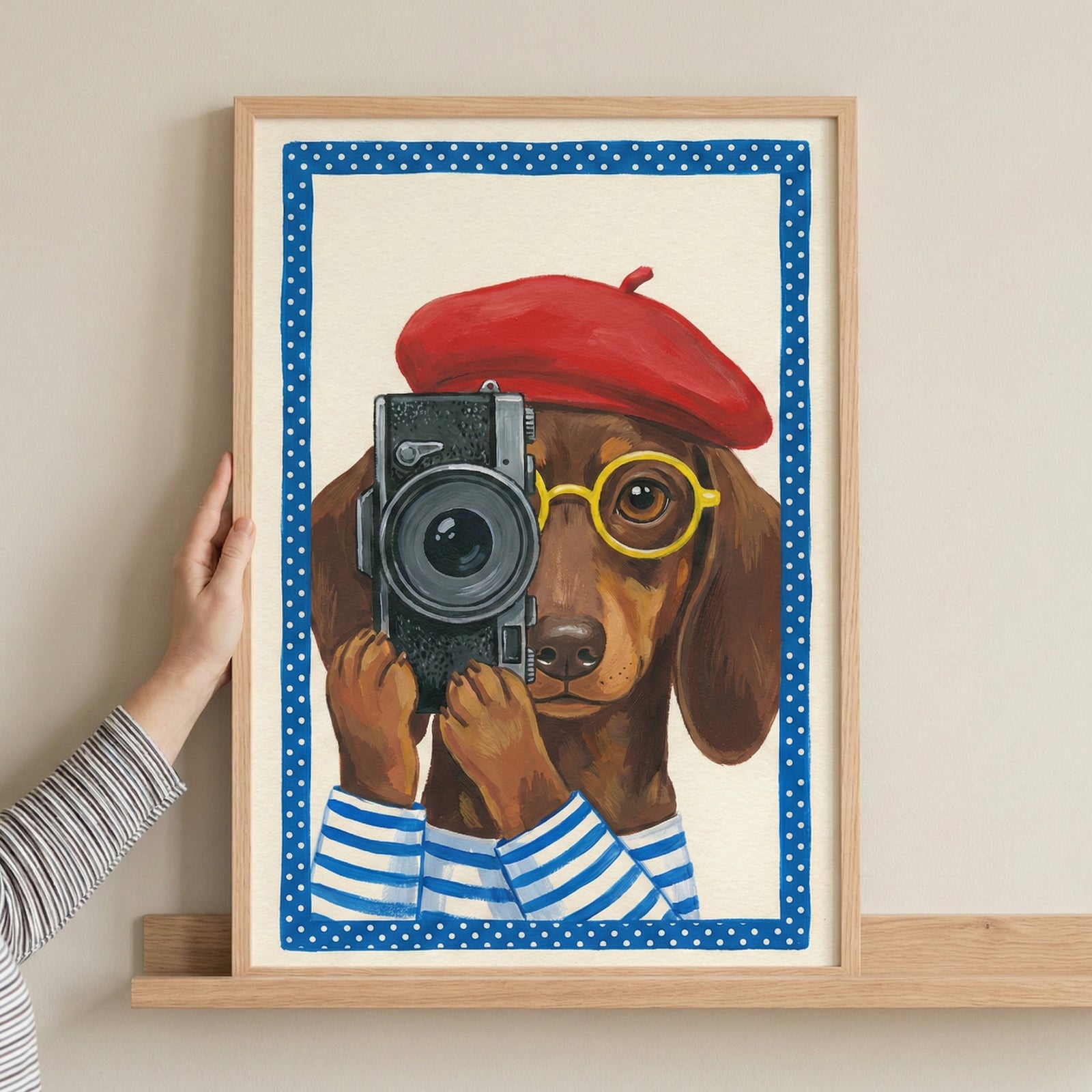 A whimsical illustration of a brown dachshund wearing a red beret, round yellow glasses, and a blue and white striped shirt. The dog holds a vintage black camera up to its eye, as if taking a photograph. The central image is surrounded by a blue border with white polka dots and is presented in a light wooden frame being held by a person's hand.