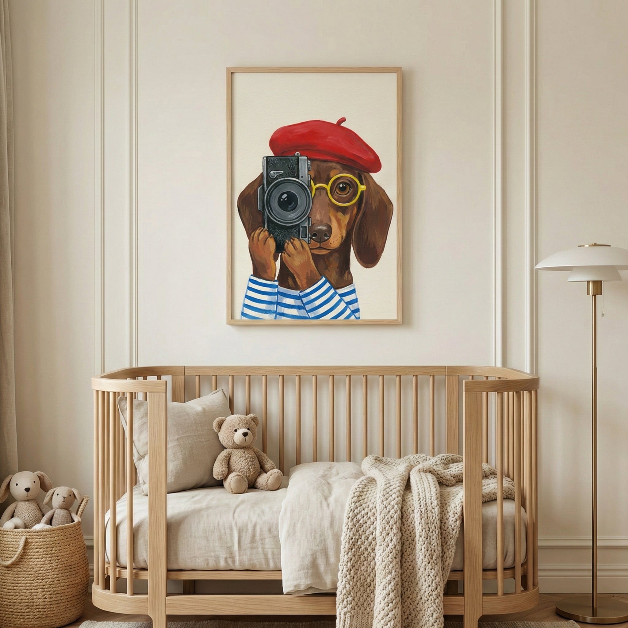 A nursery with neutral-toned walls and light wood furniture. Centered on the wall is a framed illustration of a brown dachshund wearing a red beret, yellow glasses, and a blue-and-white striped shirt, holding a black vintage camera. Below the framed artwork is a curved wooden crib containing a light brown teddy bear, a pillow, and a chunky knit blanket. To the left, a woven basket holds plush bunny toys, and to the right stands a minimalist floor lamp.