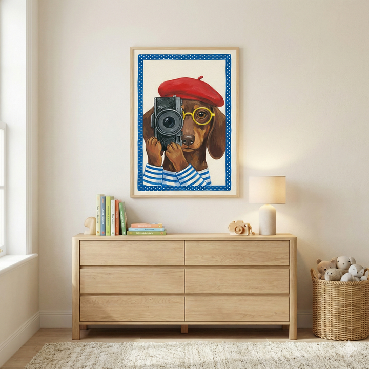 A nursery with neutral-toned walls and light wood furniture. Centered on the wall is a framed illustration of a brown dachshund wearing a red beret, yellow glasses, and a blue-and-white striped shirt, holding a black vintage camera. Below the framed artwork is a curved wooden crib containing a light brown teddy bear, a pillow, and a chunky knit blanket. To the left, a woven basket holds plush bunny toys, and to the right stands a minimalist floor lamp.