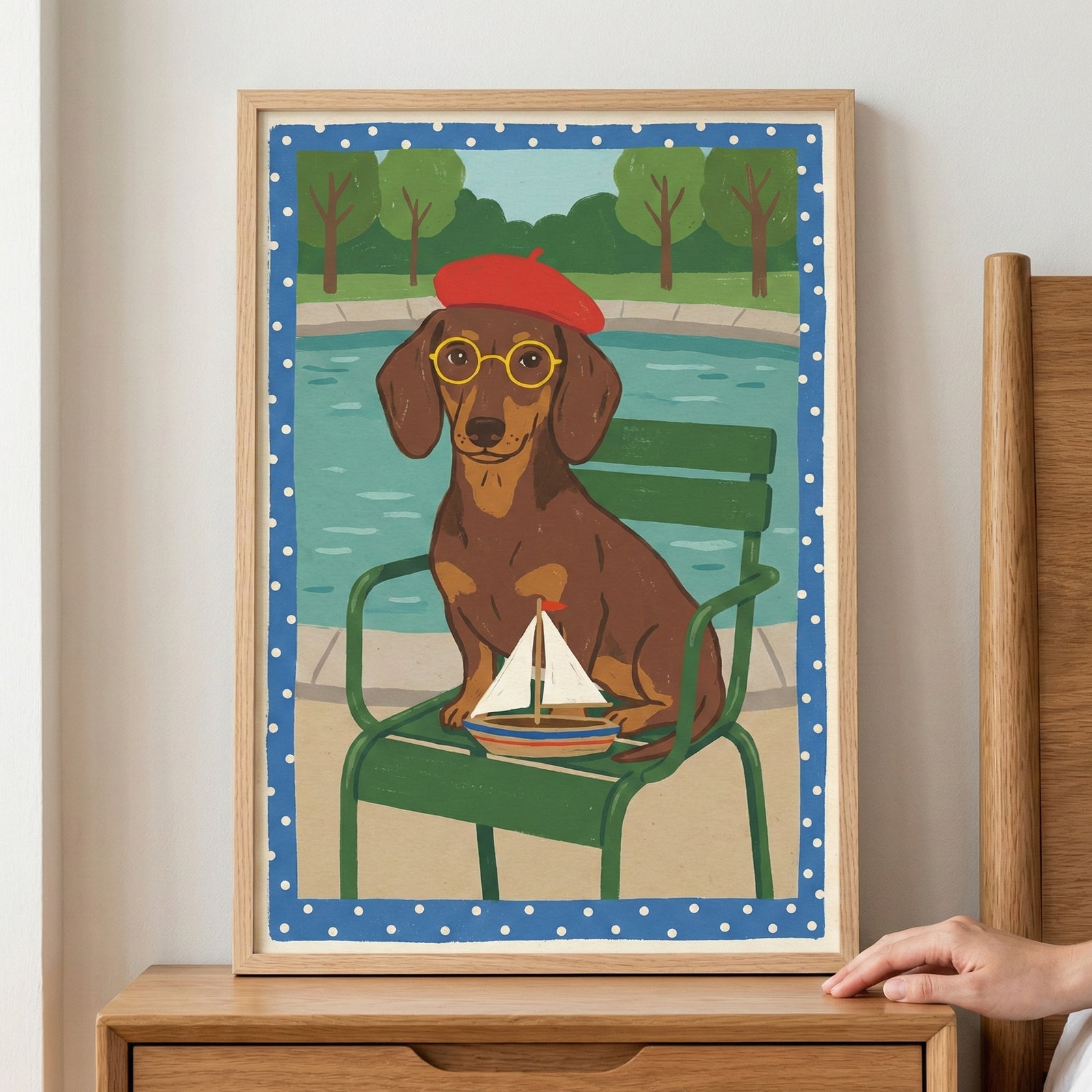 A framed illustration of a brown dachshund wearing a red beret and round yellow glasses. The dog sits on a green metal chair in front of a blue body of water. A small toy sailboat rests on the chair near the dog's paws. The background features stylized green trees. The image is bordered by a blue frame with white polka dots and is set in a light wood frame resting on a wooden bedside table.