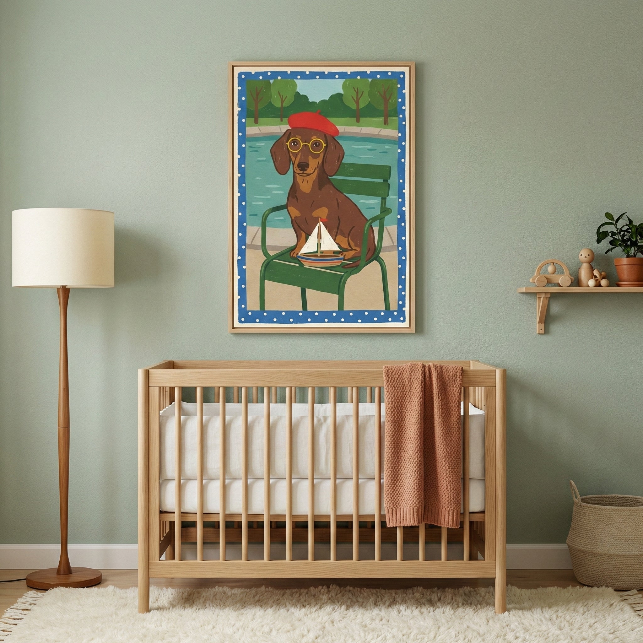 A framed illustration of a dachshund wearing a red beret and yellow glasses, sitting in a green chair and holding a toy sailboat. The artwork hangs on a sage green wall in a nursery, centered above a light wood baby crib. To the left is a tall floor lamp with a neutral shade, and to the right, a small wooden shelf holds minimalist toys and a small plant. A plush white rug covers the floor.