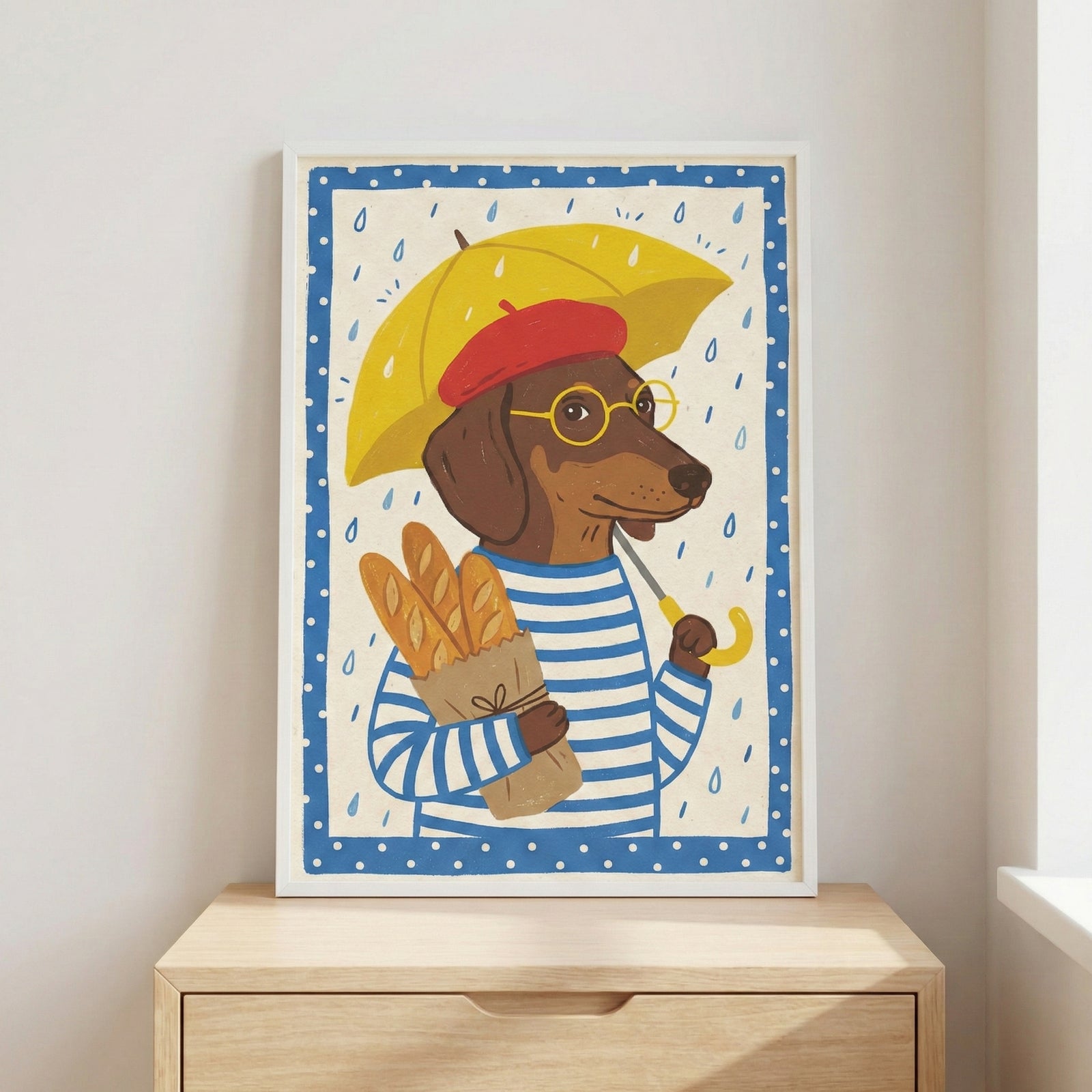 A framed illustration of a brown dachshund dressed in a blue and white striped shirt, a red beret, and yellow round glasses. The dog holds a yellow umbrella to shield itself from the rain and carries a brown paper bag with three baguettes. The artwork features a blue border with white polka dots and is placed on a light-colored wooden cabinet against a white wall.