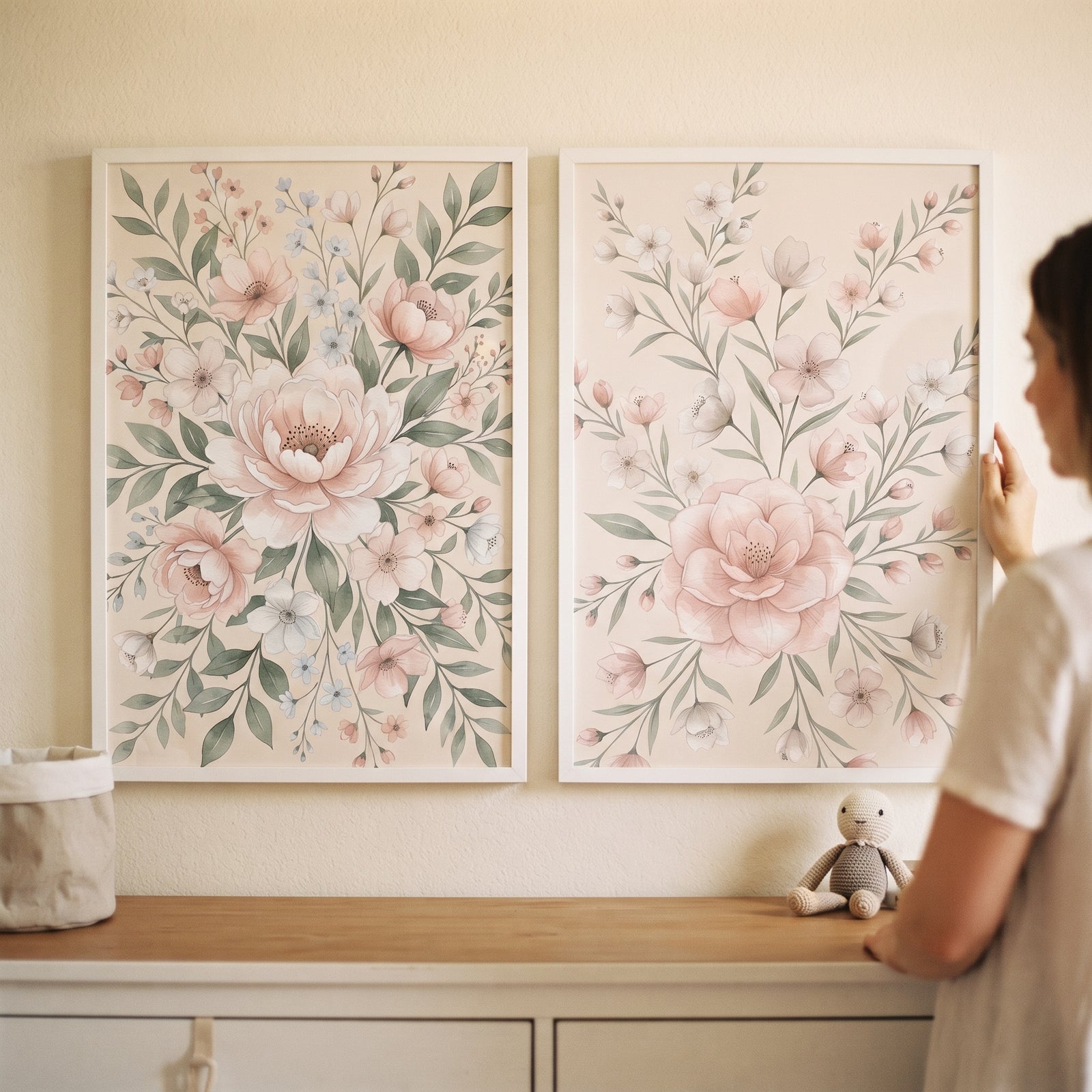 A person hangs a dainty blush floral wall art set of 2 above a wooden dresser with a small plush toy.