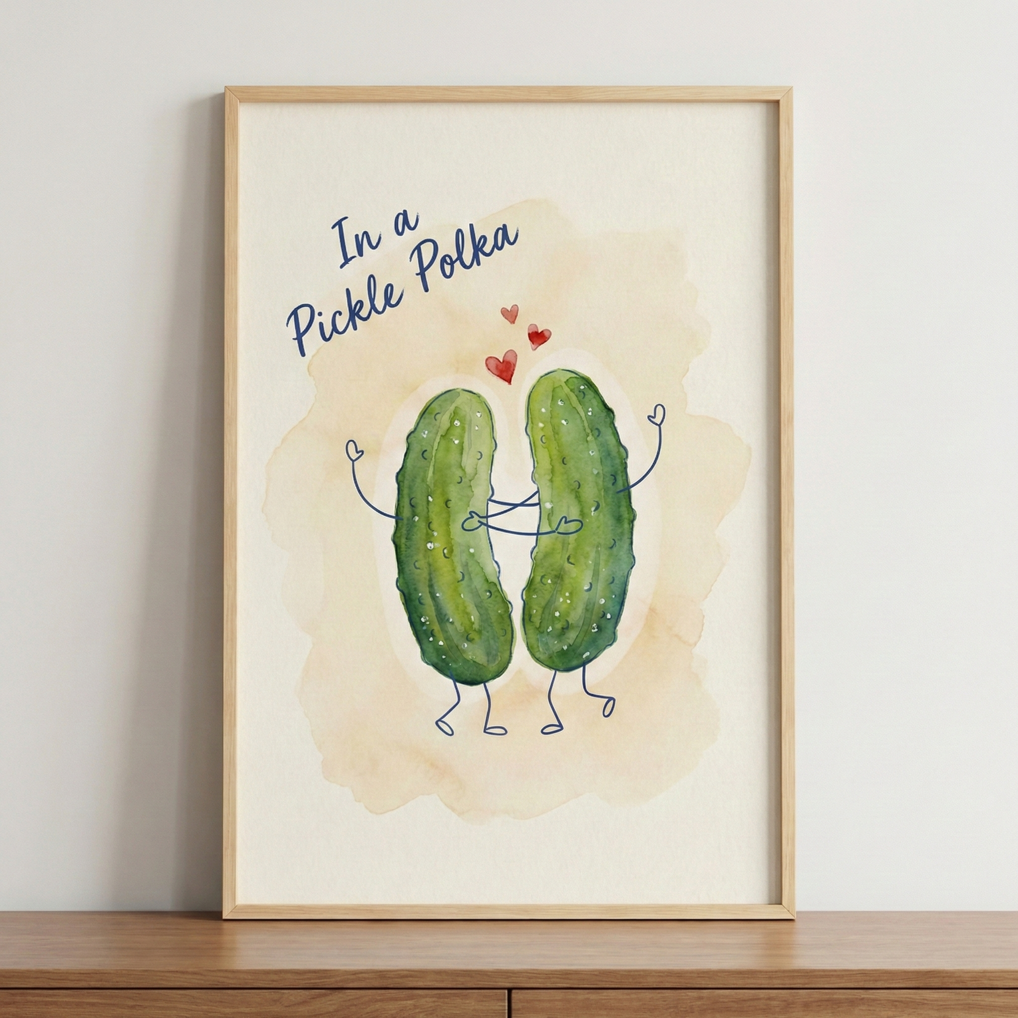 A framed watercolor illustration of two whimsical green pickles with simple stick-figure arms and legs, shown dancing or embracing. Three small red hearts float in the air between the two characters. Above them, the phrase "In a Pickle Polka" is written in a blue cursive font. The background of the artwork features a soft, beige watercolor wash. The piece is held in a light-toned wooden frame, which is sitting on a wooden surface against a plain white wall.