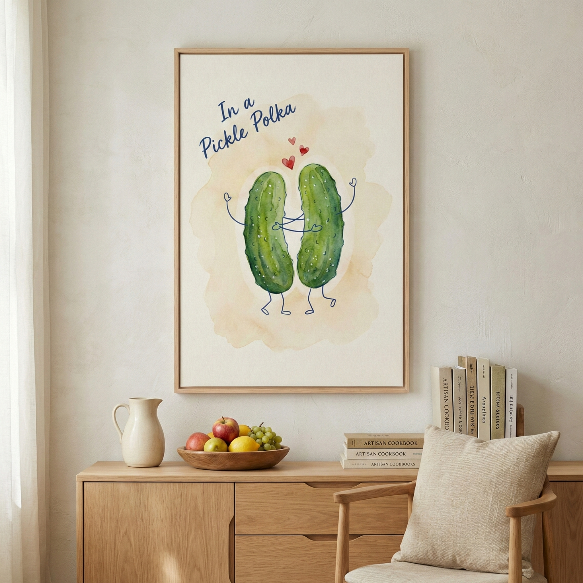 An interior scene shows a framed illustration of two cartoon pickles with thin, stick-like arms and legs, appearing to dance or embrace. Three small red hearts float above them, and the text 'In a Pickle Polka' is written in blue cursive at the top. The illustration features a soft, light-brown watercolor wash as a background. The artwork is held in a light-colored wooden frame and hangs on a cream-colored wall above a light wood console table. 