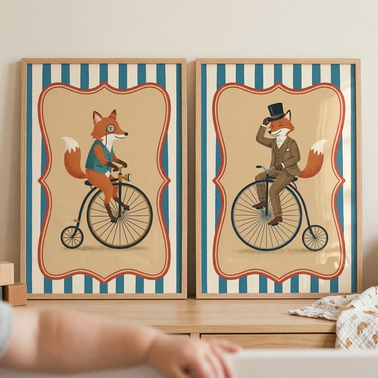 Two vertical wooden-framed illustrations sit on a light-colored wooden surface. Both artworks feature a whimsical orange fox riding a vintage-style bicycle with a large front wheel and a small rear wheel. 

