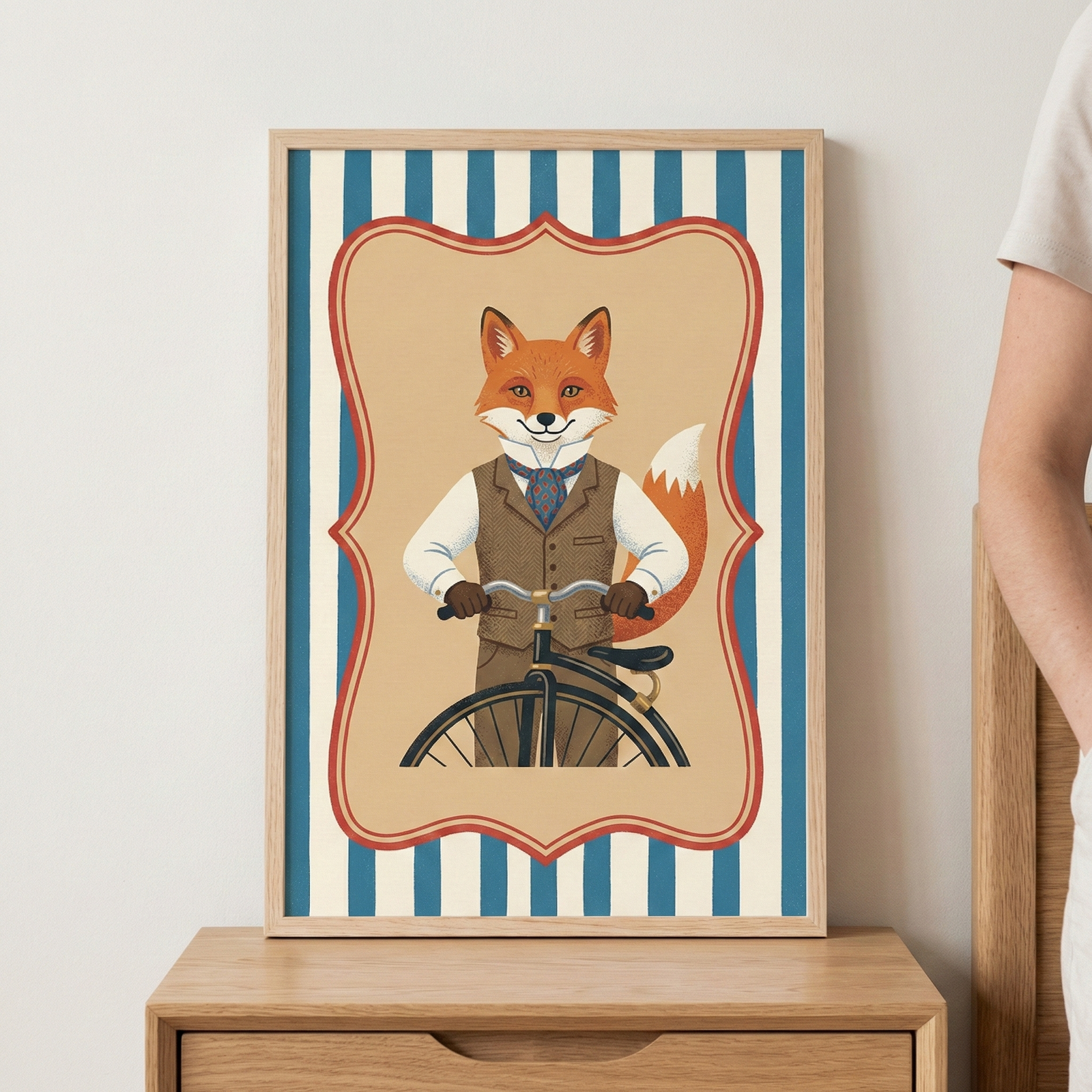 A framed illustration of an anthropomorphic fox dressed in vintage attire, including a brown vest and a blue patterned cravat. The fox is holding the handlebars of a vintage-style bicycle. The central image is set within a decorative scalloped border against a background of vertical blue and cream stripes. The frame rests on a light-colored wooden surface.