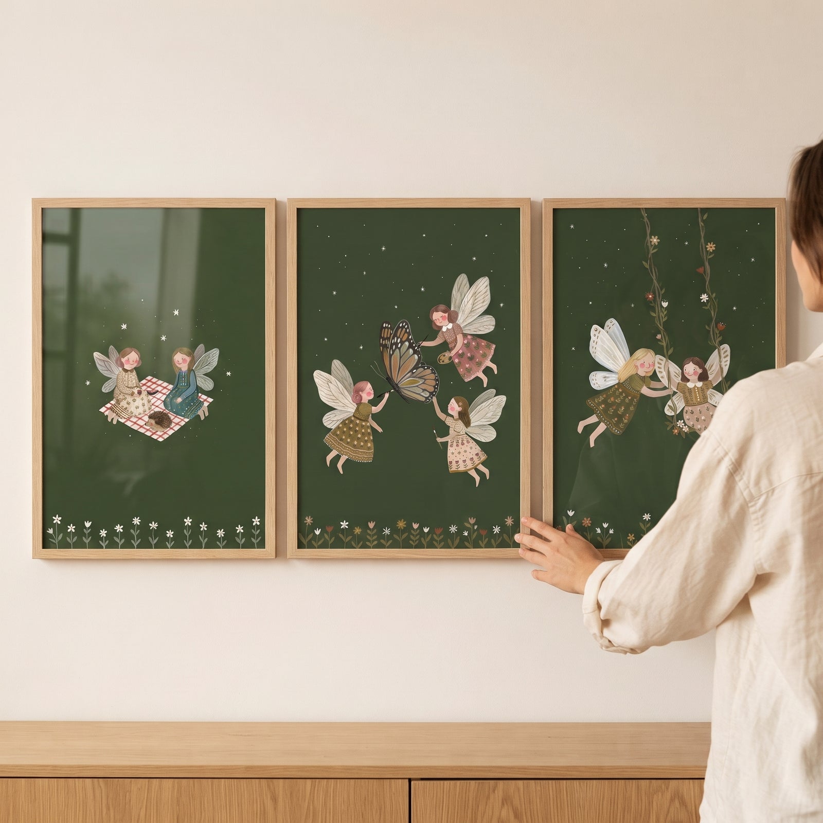 A person hangs a dark green woodland fairies wall art set of 3 in wooden frames above a cabinet.