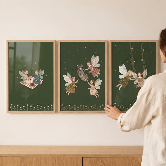 A person hangs a dark green woodland fairies wall art set of 3 in wooden frames above a cabinet.
