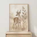 A vertical framed illustration in a light wood frame features a young deer with small antlers standing in a field of tall grass. The artwork is done in a soft, watercolor style using a neutral color palette of beige, tan, and brown. The deer has white spots on its back and gazes to the side. A slender, leafy branch stands tall to the right of the deer. The framed art sits on a light wooden dresser against a plain, off-white wall.