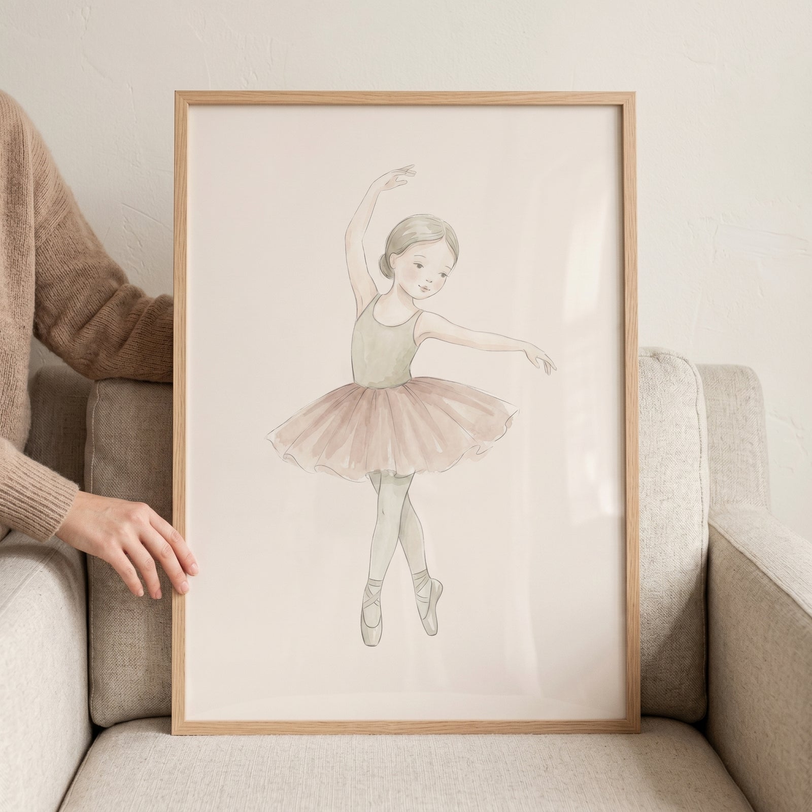Framed watercolor illustration of a young ballerina in a soft pink tutu and sage green leotard, held against a grey chair.