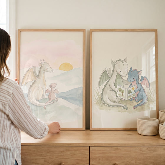 delicate watercolor baby dragons wall art set of 2 with white, pink, and blue dragons at sunset and reading a book.