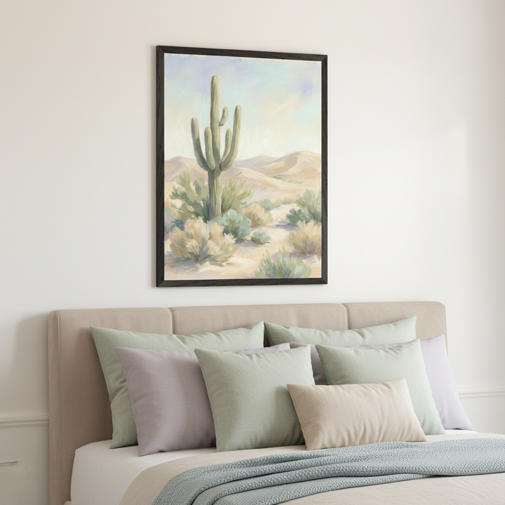 Modern bedroom with a bed featuring a beige headboard and multiple pastel pillows. A framed painting of a desert landscape with a saguaro cactus, shrubs, and sand dunes hangs centrally on the white wall above the bed.