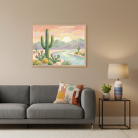 Living room with a grey modular sofa. Centered above the sofa, a framed painting depicts a vibrant desert landscape with a large saguaro cactus, blooming cacti, a river, mountains, and a sunset, in a painterly style. A colorful throw pillow rests on the sofa. A side table with a lamp and small cactus is to the right.