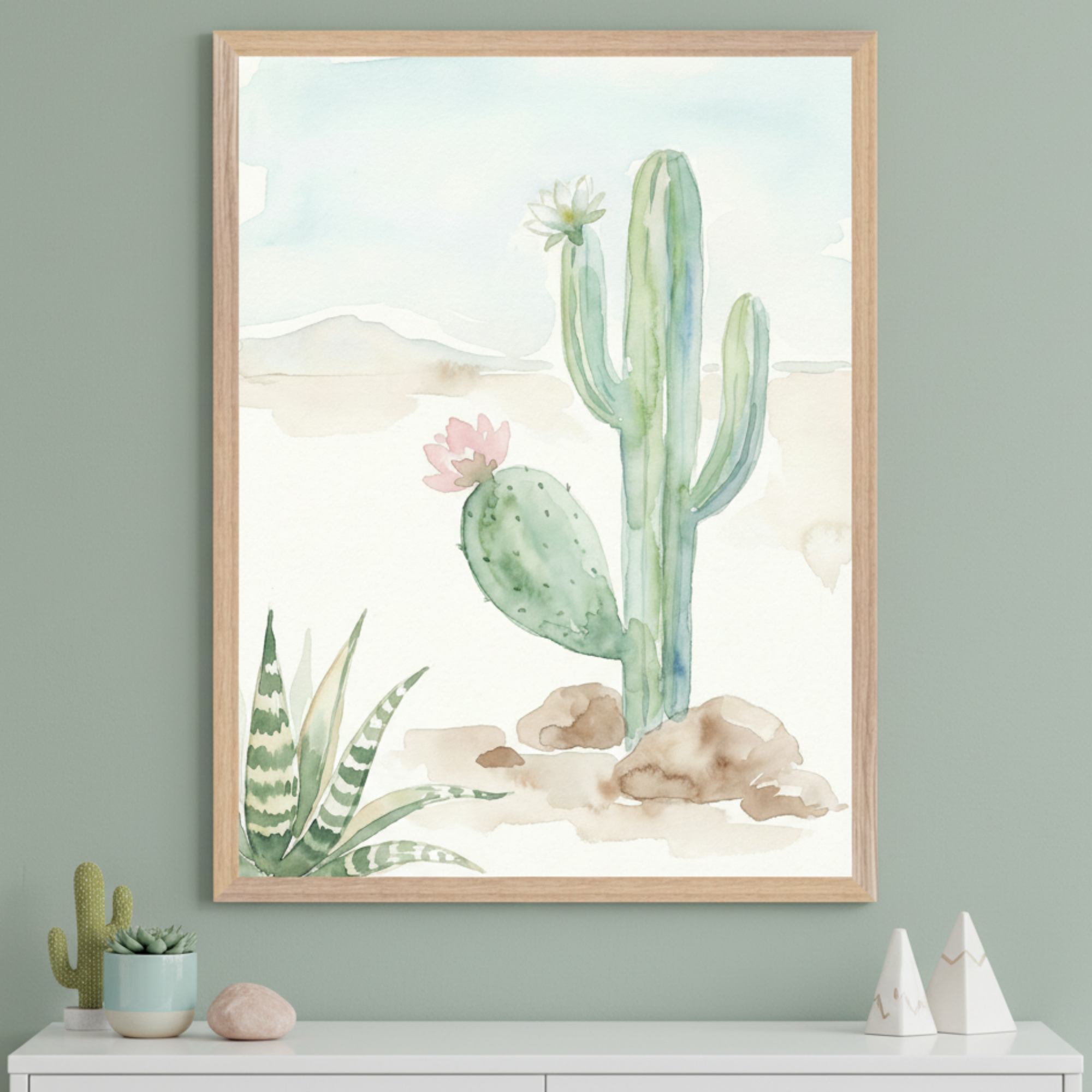 Modern room with a sage green wall and white dresser. A large framed watercolor painting of desert cacti, including a saguaro with a white flower and a prickly pear with a pink flower, hangs above the dresser. Small plants and decorative items are on the dresser.