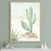 Modern room with a sage green wall and white dresser. A large framed watercolor painting of desert cacti, including a saguaro with a white flower and a prickly pear with a pink flower, hangs above the dresser. Small plants and decorative items are on the dresser.