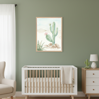 Modern nursery with a sage green wall. A framed watercolor desert scene with a tall cactus, flowering cactus, and succulent hangs above a white crib with wood spindles. A beige armchair is on the left, and a wood dresser with a lamp and cactus decor is on the right.