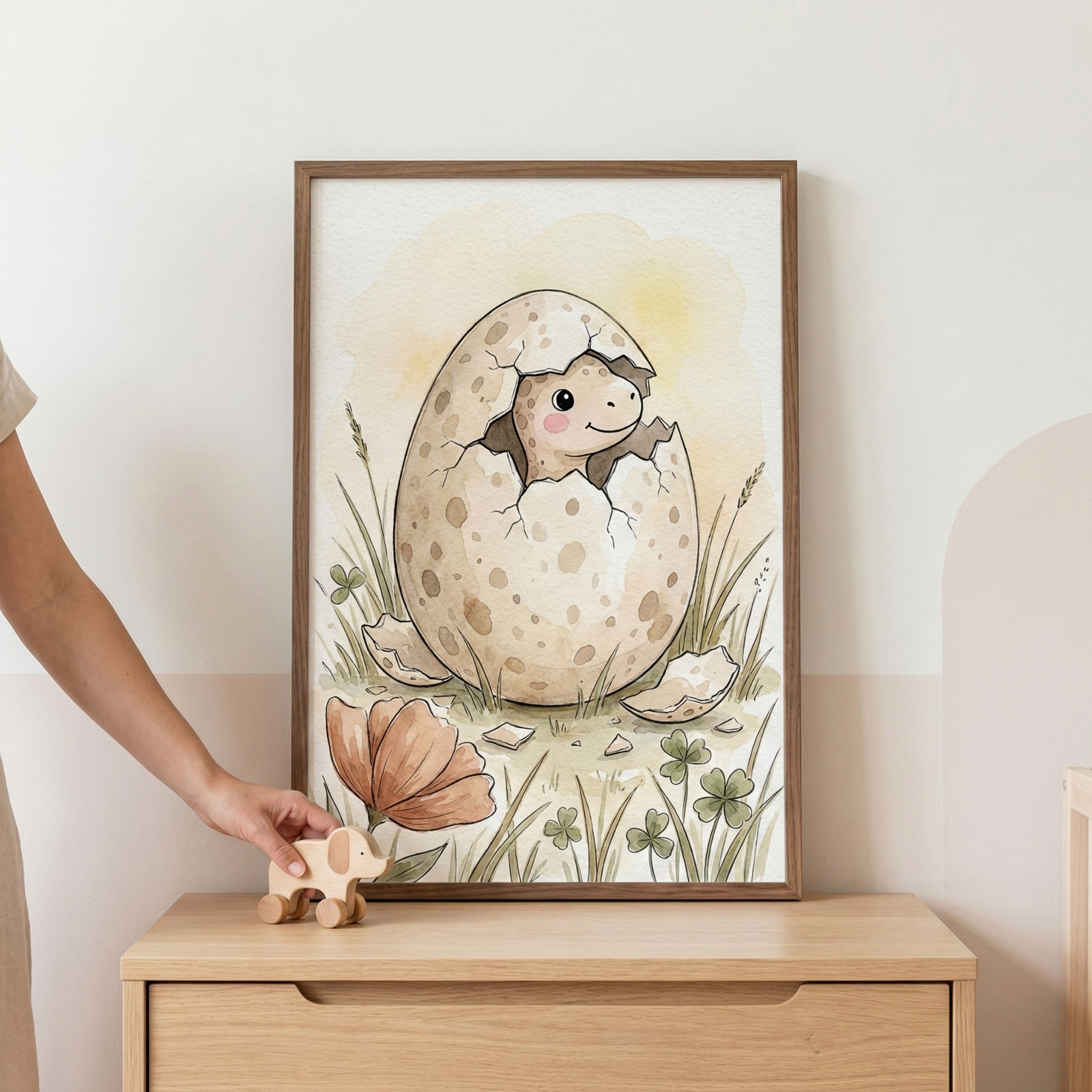 A framed watercolor illustration of a smiling baby dinosaur hatching from a spotted egg, surrounded by green grass, clovers, and a large peach-colored flower. The frame is displayed on a light wood dresser, and a hand is visible holding a small wooden animal toy on wheels next to it.
