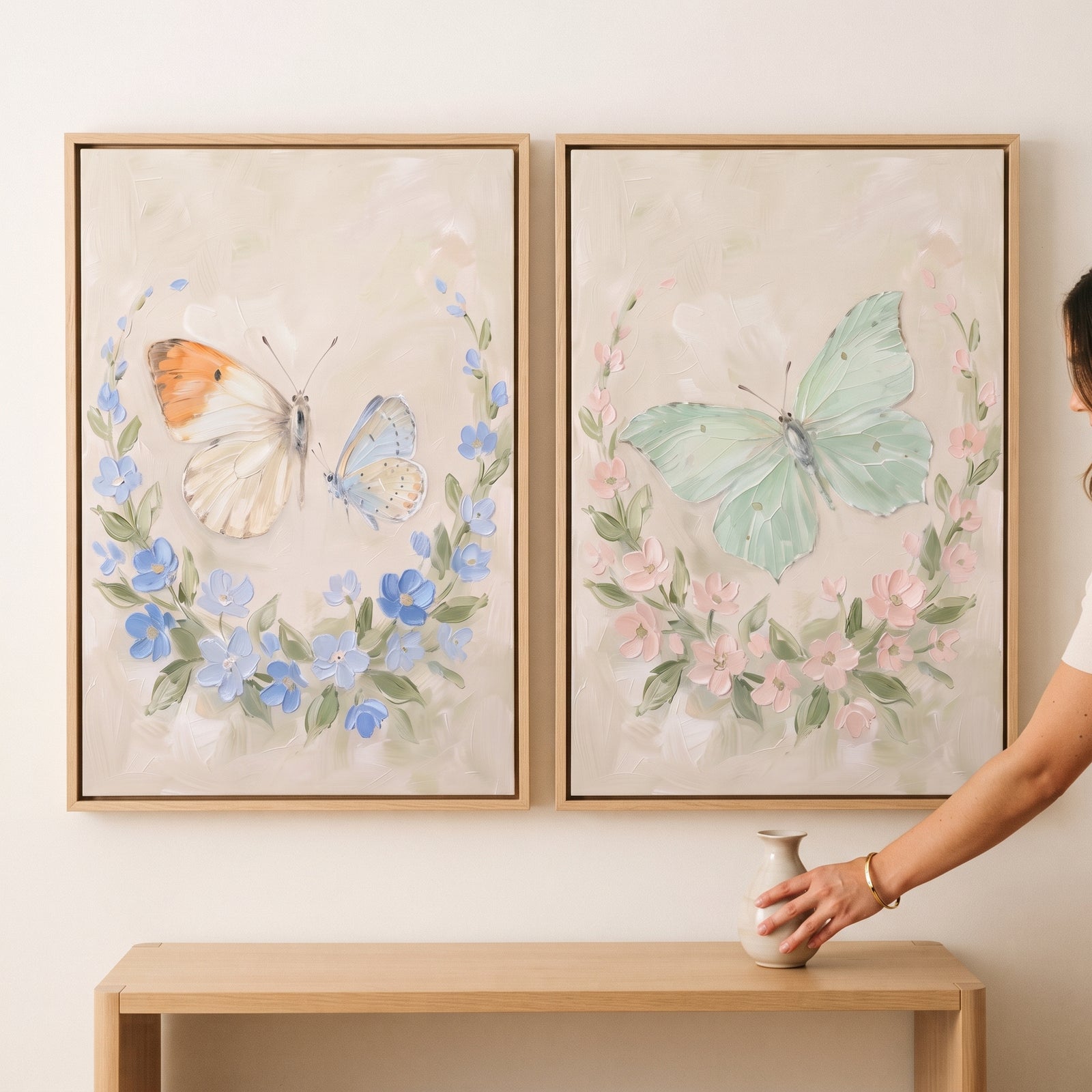 Distressed neutral floral butterfly print set of 2 hangs above a wooden table with a hand adjusting a vase.