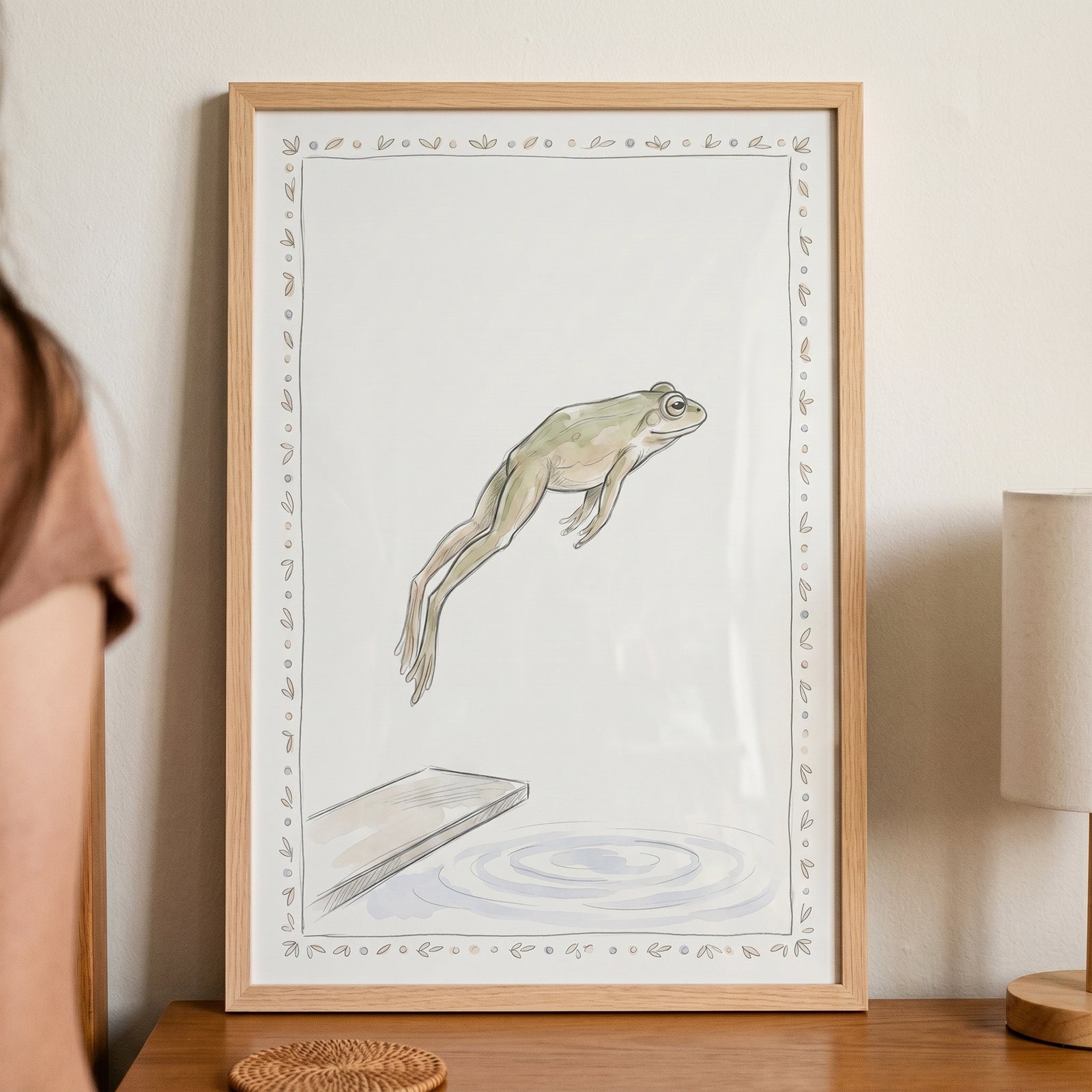 Diver green frog wall art in a light wood frame shows a frog leaping from a diving board into a pool of water.
