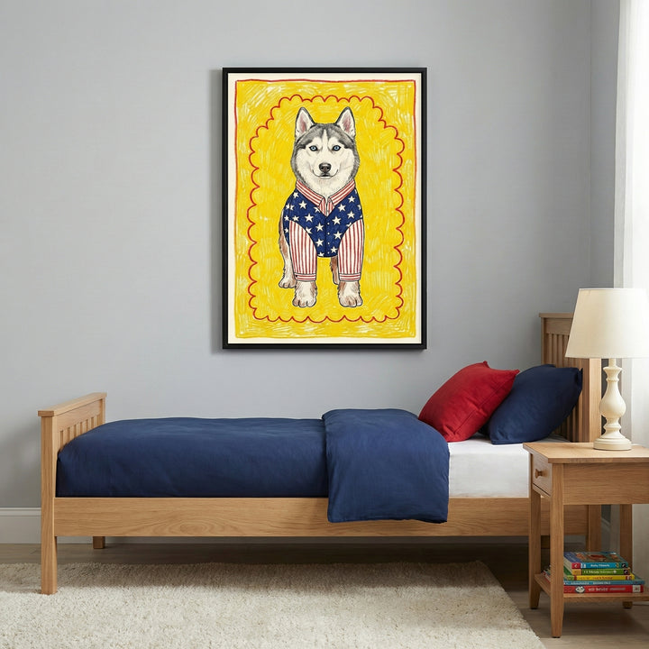 A framed whimsical illustration of a husky wearing an American flag-patterned shirt hangs above a wooden twin bed with navy blue and red bedding. The room features gray walls, a white shag rug, and a simple wooden nightstand with a lamp and a stack of books.