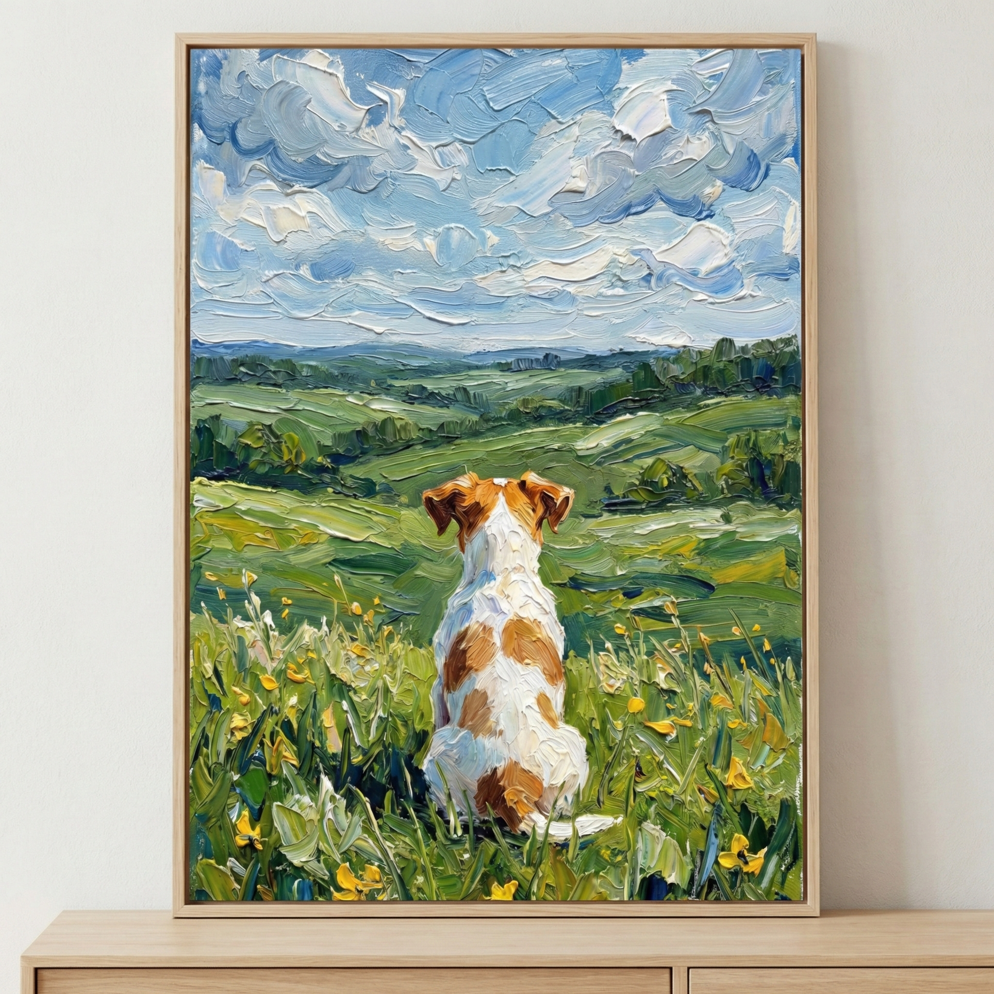 A small white and tan dog sits in a field of green grass and yellow wildflowers, seen from behind. The dog gazes out over a scenic vista of rolling green hills. Above, a vast blue sky is filled with thick, textured white clouds. The entire scene is rendered with heavy, visible strokes of color, creating a rich, tactile texture. This artwork is presented in a light wooden frame resting on a wooden surface against a neutral background.