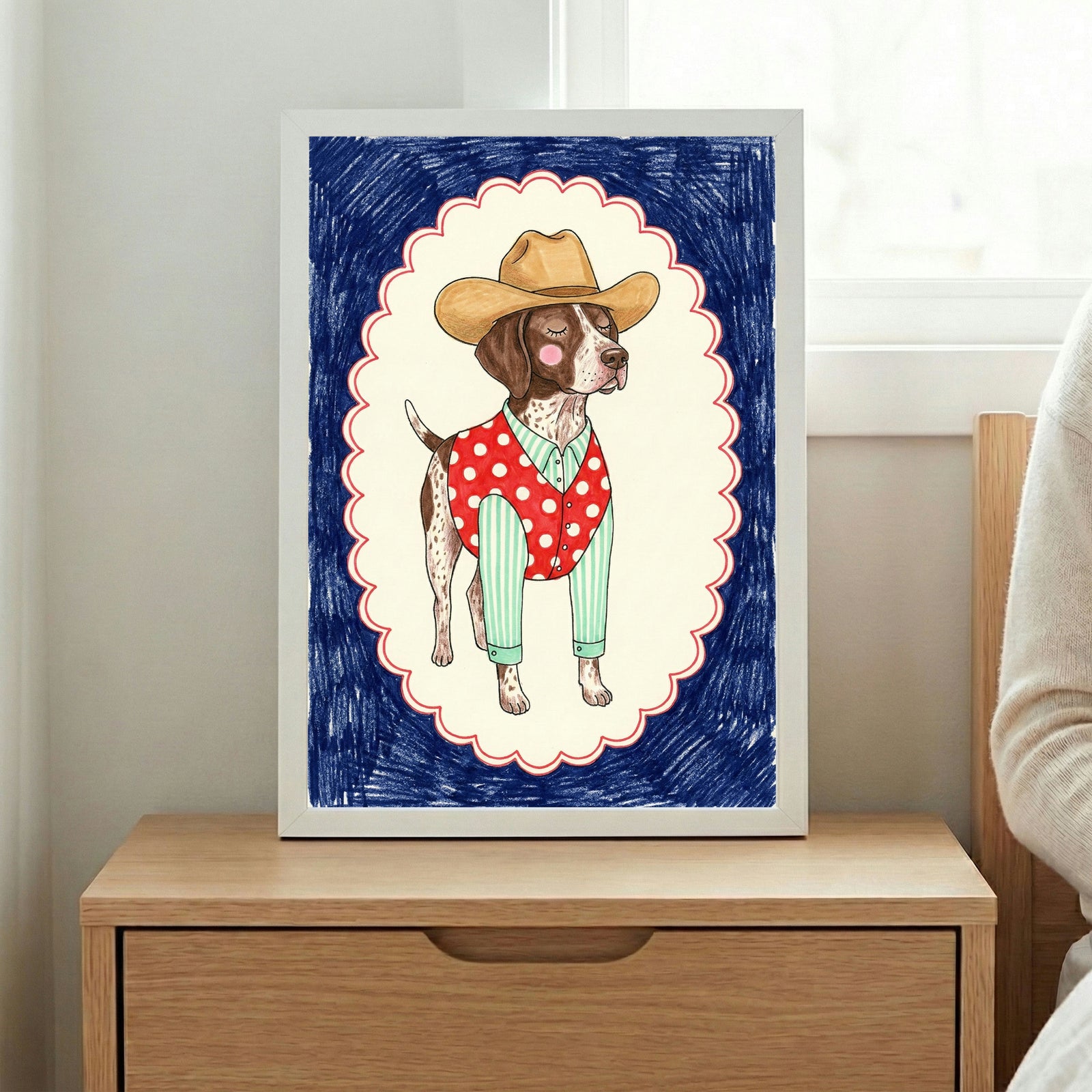 Whimsical illustration of a dog dressed as a cowboy, wearing a tan hat, green striped shirt, and red polka-dotted vest. The dog stands proudly within a scalloped white frame outlined in red, set against a dark blue scribbled background. The framed art is placed on a wooden nightstand next to a window.