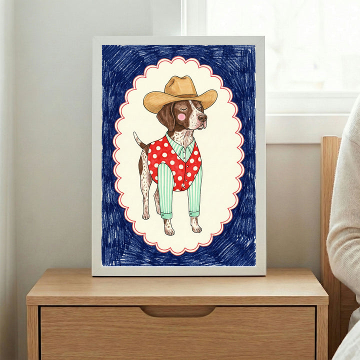 Whimsical illustration of a dog dressed as a cowboy, wearing a tan hat, green striped shirt, and red polka-dotted vest. The dog stands proudly within a scalloped white frame outlined in red, set against a dark blue scribbled background. The framed art is placed on a wooden nightstand next to a window.