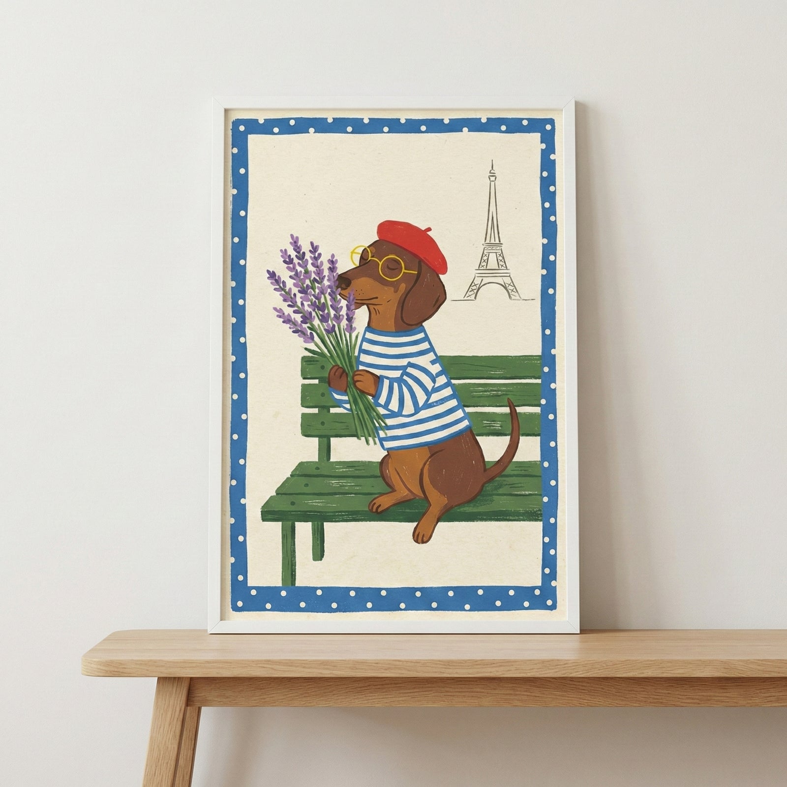 An illustration in a white frame sits on a wooden table against a neutral wall. The artwork depicts a brown dachshund wearing a red beret, round glasses, and a blue-and-white striped shirt. The dog is sitting on a green park bench, holding a large bunch of purple lavender flowers. In the background, there is a simple line drawing of a famous landmark tower. The central image is framed by a wide blue border with white polka dots.