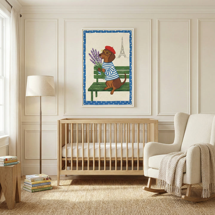 A neutral-toned nursery features a light wood crib centered against a paneled wall. Above the crib hangs a framed illustration of a small brown dog wearing a red beret and a blue-and-white striped shirt, sitting on a green park bench and holding a bunch of lavender. The illustration includes a simple line drawing of a famous landmark tower in the background and is framed by a blue border with white polka dots. To the right of the crib is a cream-colored rocking chair with a knit throw blanket draped over it