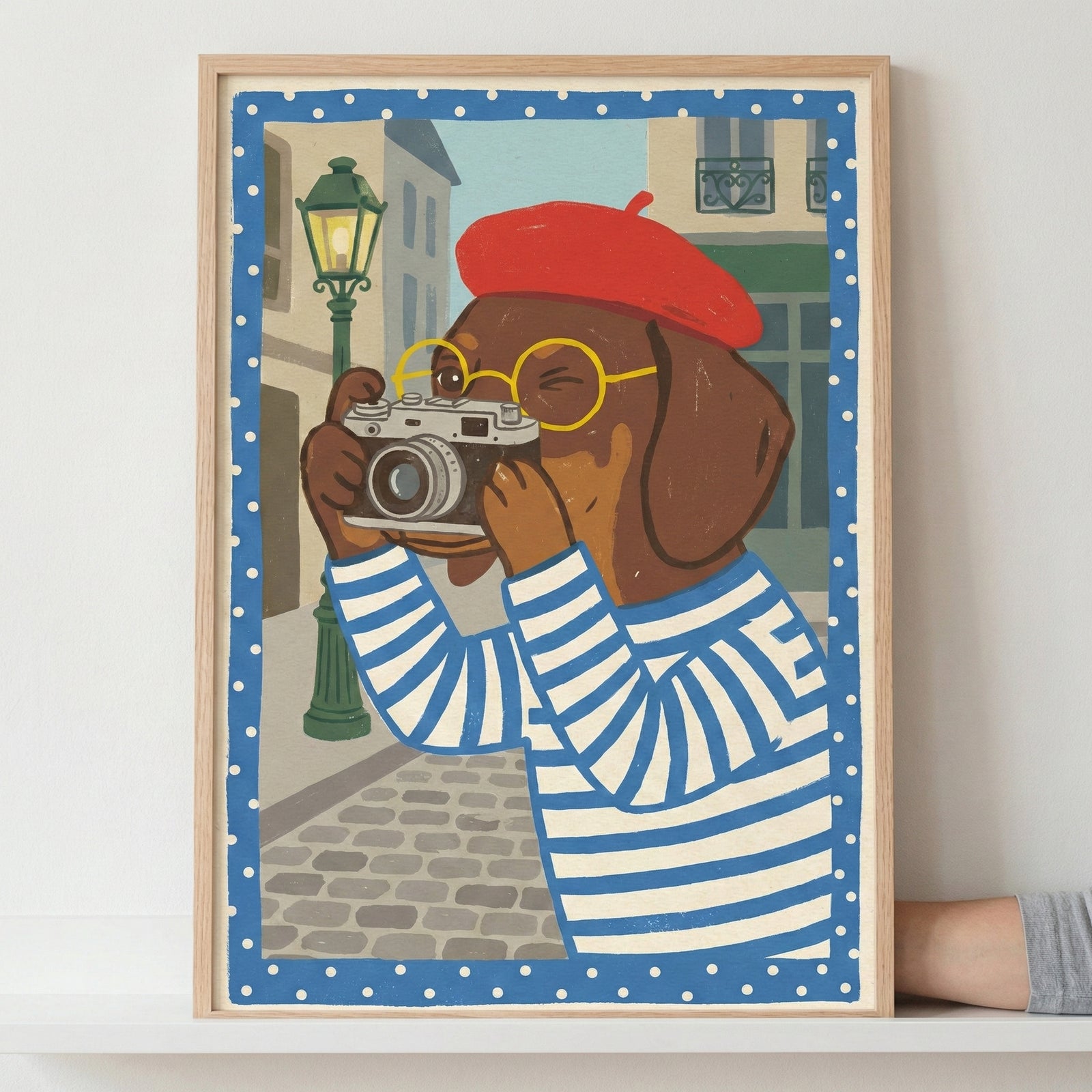 A framed illustration of an anthropomorphic brown dog with long ears, dressed in a red beret, yellow round-framed glasses, and a blue and white striped shirt. The dog is holding a vintage-style camera to its eye, appearing to take a photograph. The background features a cobblestone street with a green lamppost and city buildings. The image has a blue border with white polka dots and is set within a light-colored wooden frame, which is being held by a hand against a white wall.