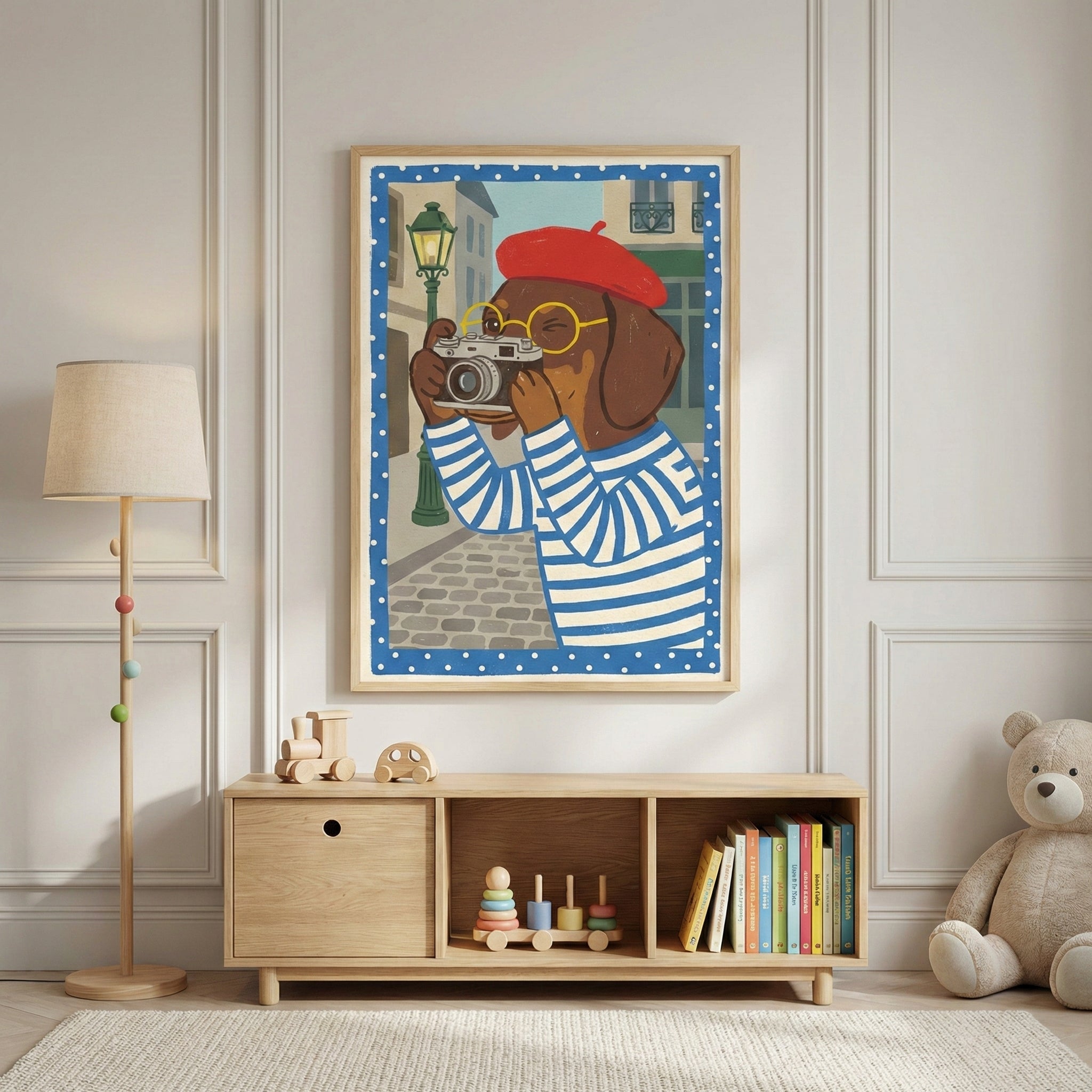 A framed illustration of a brown dog wearing a red beret, yellow glasses, and a blue-and-white striped shirt hangs on a light-colored paneled wall. In the artwork, the dog holds a vintage-style camera against a backdrop of a European street. Below the frame sits a light wood storage unit with three compartments containing wooden toys and a collection of colorful books. To the left is a tall wooden floor lamp with a beige shade, and to the right, a large plush teddy bear sits on the floor. The scene is set i