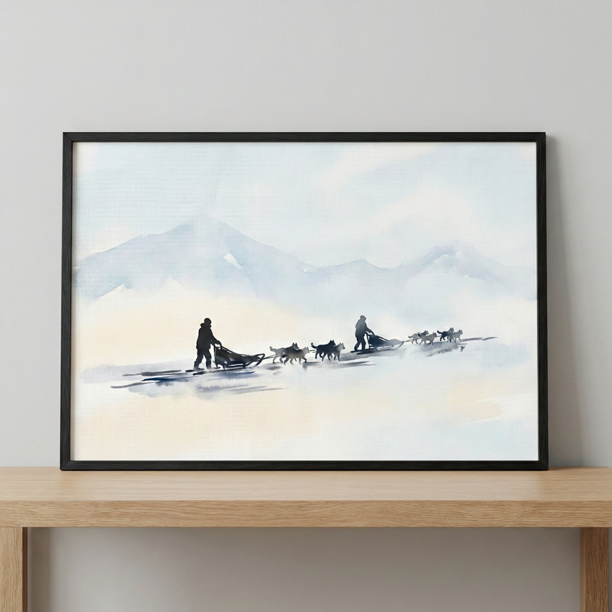 A framed horizontal piece of art depicting two dog sled teams traveling across a vast, snowy landscape. The silhouettes of the dogs and mushers are rendered in dark tones, contrasting with the soft, atmospheric washes of blue, grey, and cream that form the background of hazy mountains and a pale sky. The style is minimalist with a soft watercolor-like aesthetic.