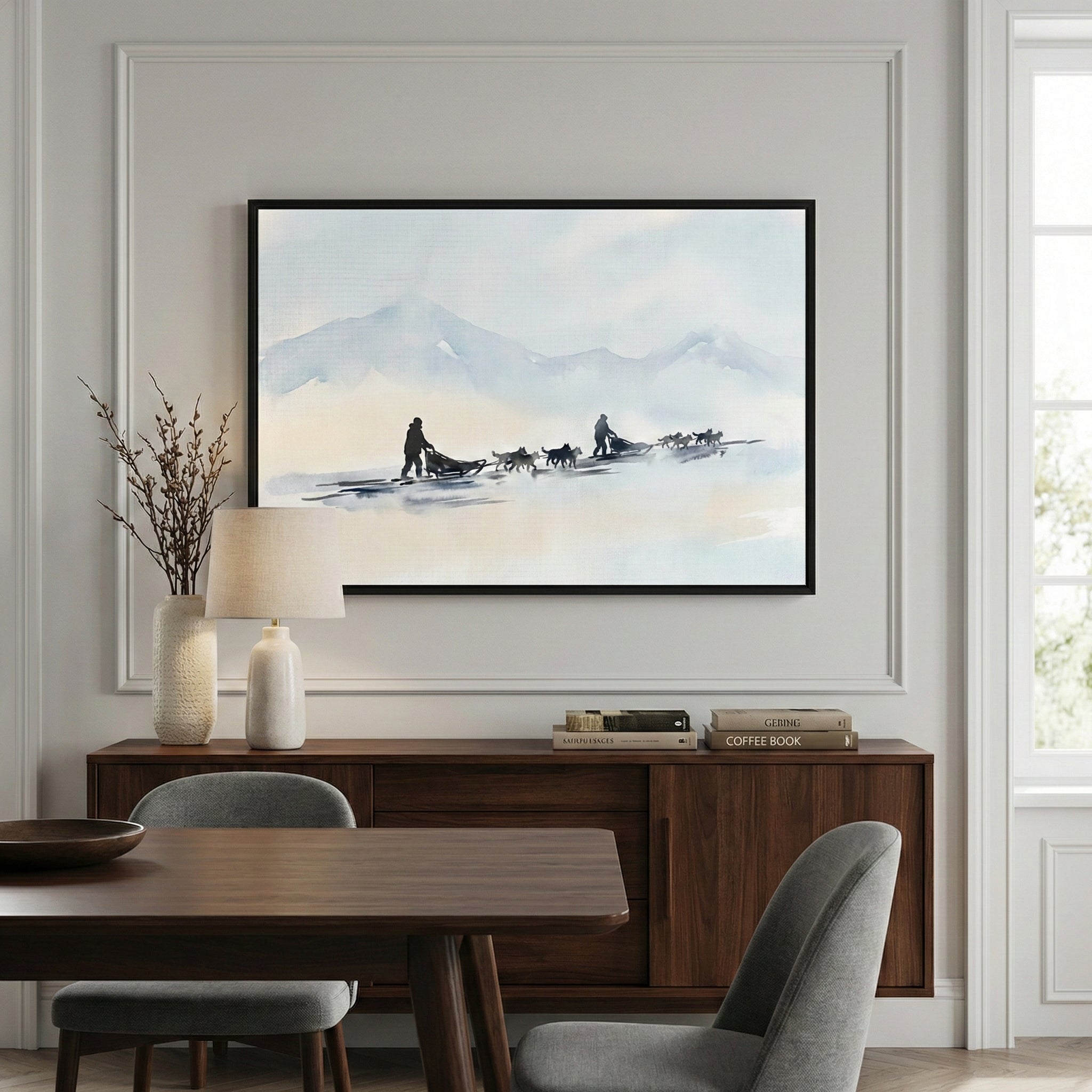 A modern dining room features a large framed artwork centered on a wall with decorative molding. The artwork shows a serene winter landscape with dog sled teams traveling across a snowy expanse toward distant blue mountains in a soft, washed-out style. Below the frame, a dark wood sideboard holds a textured white vase with dried twigs, a table lamp with a cream shade, and a few books. In the foreground, a dark wood dining table is paired with grey upholstered chairs.