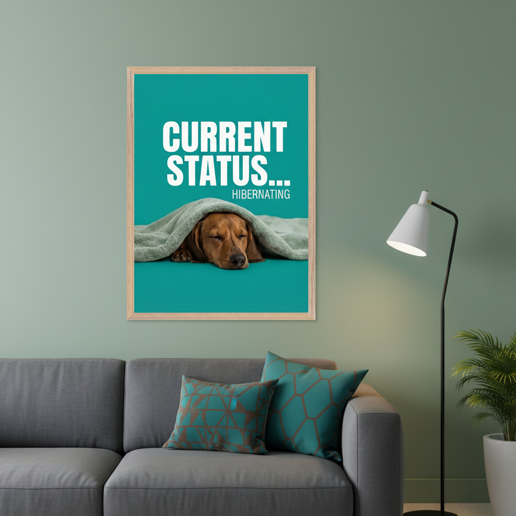 Living room with a gray sofa, two patterned pillows, and a floor lamp. Above the sofa, a framed poster features a sleeping dachshund under a blanket on a teal background with text "CURRENT STATUS... HIBERNATING".