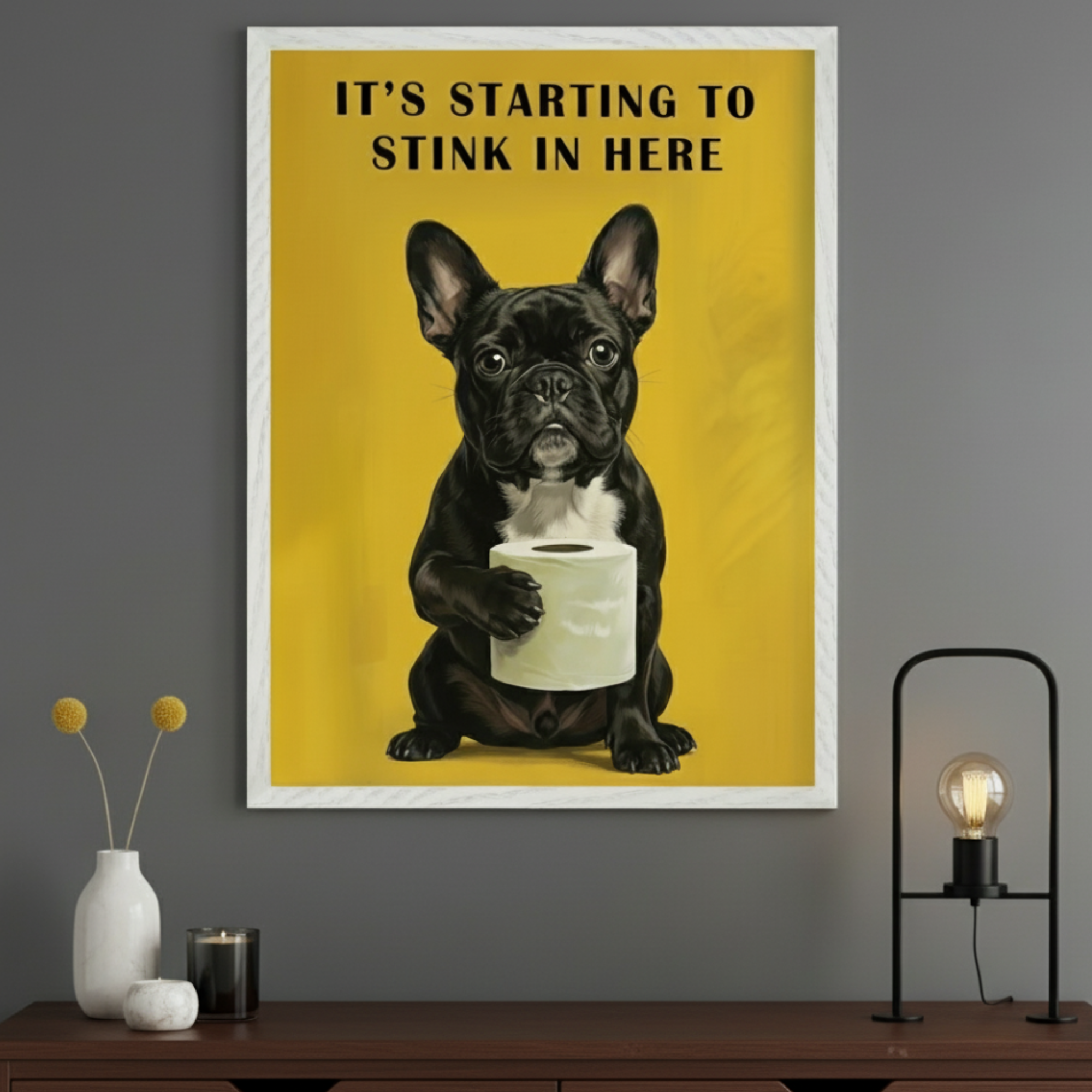 Modern room with a dark wood dresser. A framed graphic print of a black French Bulldog holding toilet paper, with text "IT'S STARTING TO STINK IN HERE" on a yellow background, hangs above the dresser. Decor on dresser includes a vase, candles, and a lamp.