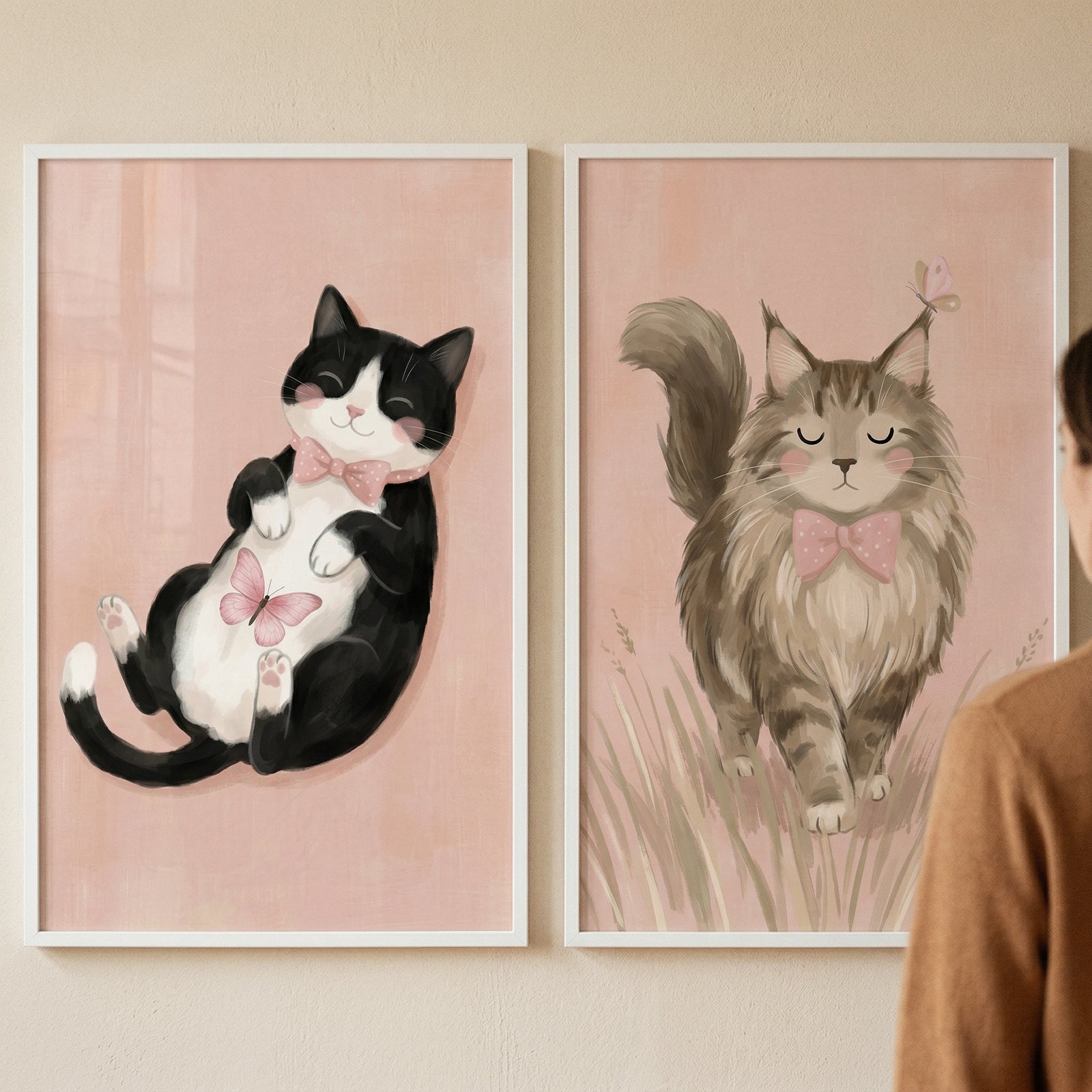 Two white-framed illustrations are displayed side-by-side on a light-toned wall. Both pieces feature a soft pink background and a whimsical depiction of a cat. 

The illustration on the left shows a black and white tuxedo cat lying playfully on its back. It has a smiling face with rosy cheeks and wears a light pink bow tie. A small pink butterfly rests on its white fur. 

