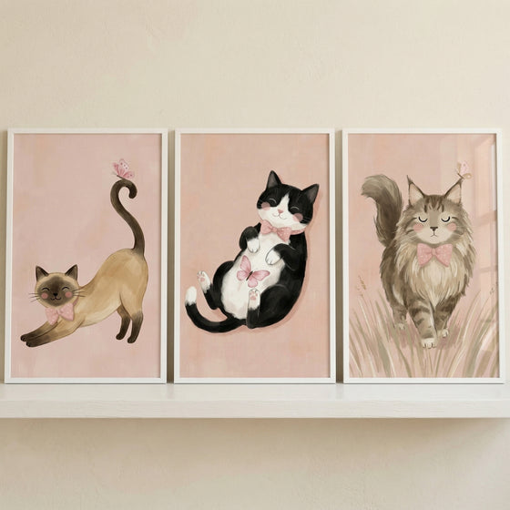 A set of three framed illustrations featuring different cats is displayed side-by-side on a white floating shelf. Each artwork is set against a soft, pale pink background.

In the first illustration on the left, a slender tan cat with dark markings on its face and paws is shown stretching, its tail curled high with a tiny pink butterfly on the tip. It wears a small pink bow tie.
