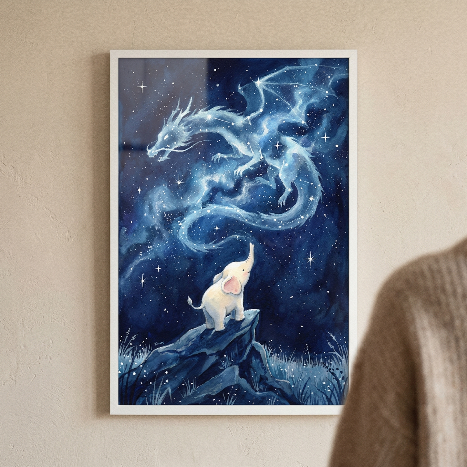 A framed piece of fantasy art hangs on a neutral-colored wall. Inside a thin white frame, the illustration depicts a small white elephant calf standing on a dark, rocky outcrop, looking up at a vast, dark blue night sky. A large, ethereal dragon, seemingly formed from stars and glowing celestial clouds, stretches across the sky above the elephant. The scene is filled with twinkling stars and soft, shimmering light. Part of a person's shoulder in a brown sweater is blurred in the foreground.