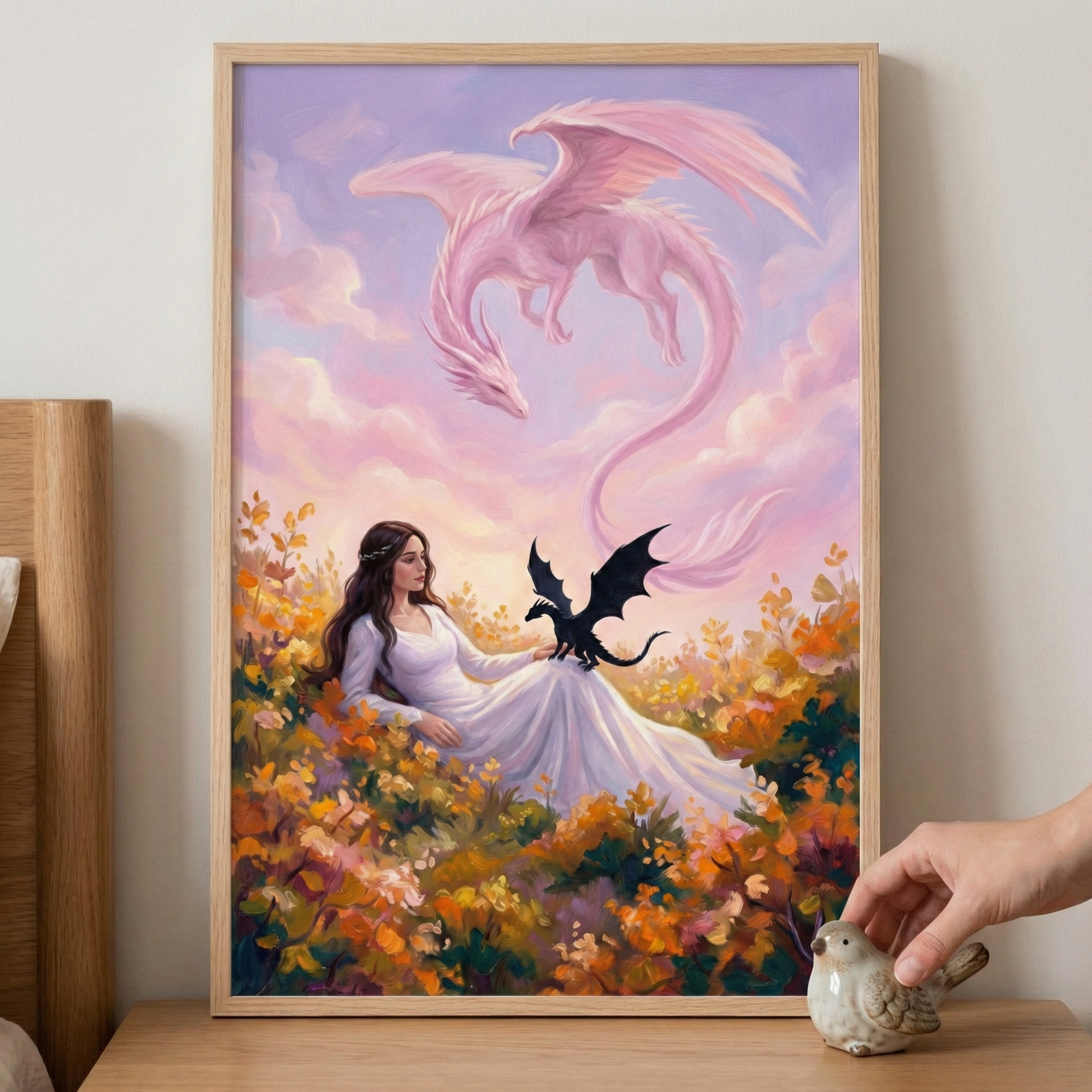 A framed fantasy illustration displays a woman in a flowing white dress sitting in a dense field of orange and yellow flowers. A small, black dragon is perched on her lap. In the soft pink and purple sky above, a large, translucent pink dragon with a long tail soars. The artwork is held in a light wood frame on a wooden surface. To the right, a person's hand reaches toward a small, light-colored ceramic bird figurine.