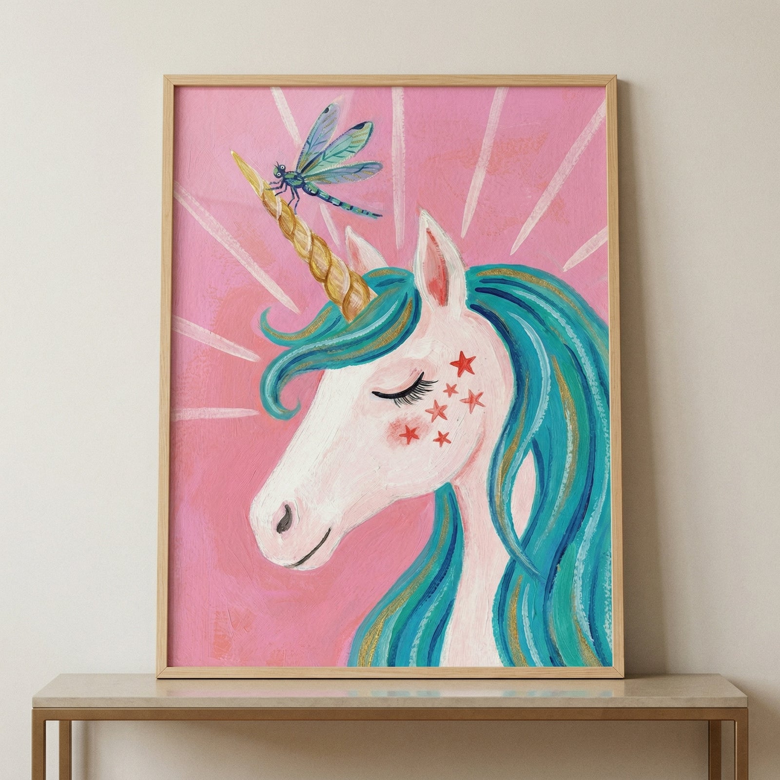 This framed illustration shows the side profile of a white unicorn with a spiraled gold horn and a flowing teal mane. A colorful dragonfly is landed on the horn, and small red stars decorate the creature's cheek. The background is a solid pink color with white rays emanating from the central figure. This creative work is enclosed in a light wood frame and displayed on a simple table against a neutral wall.