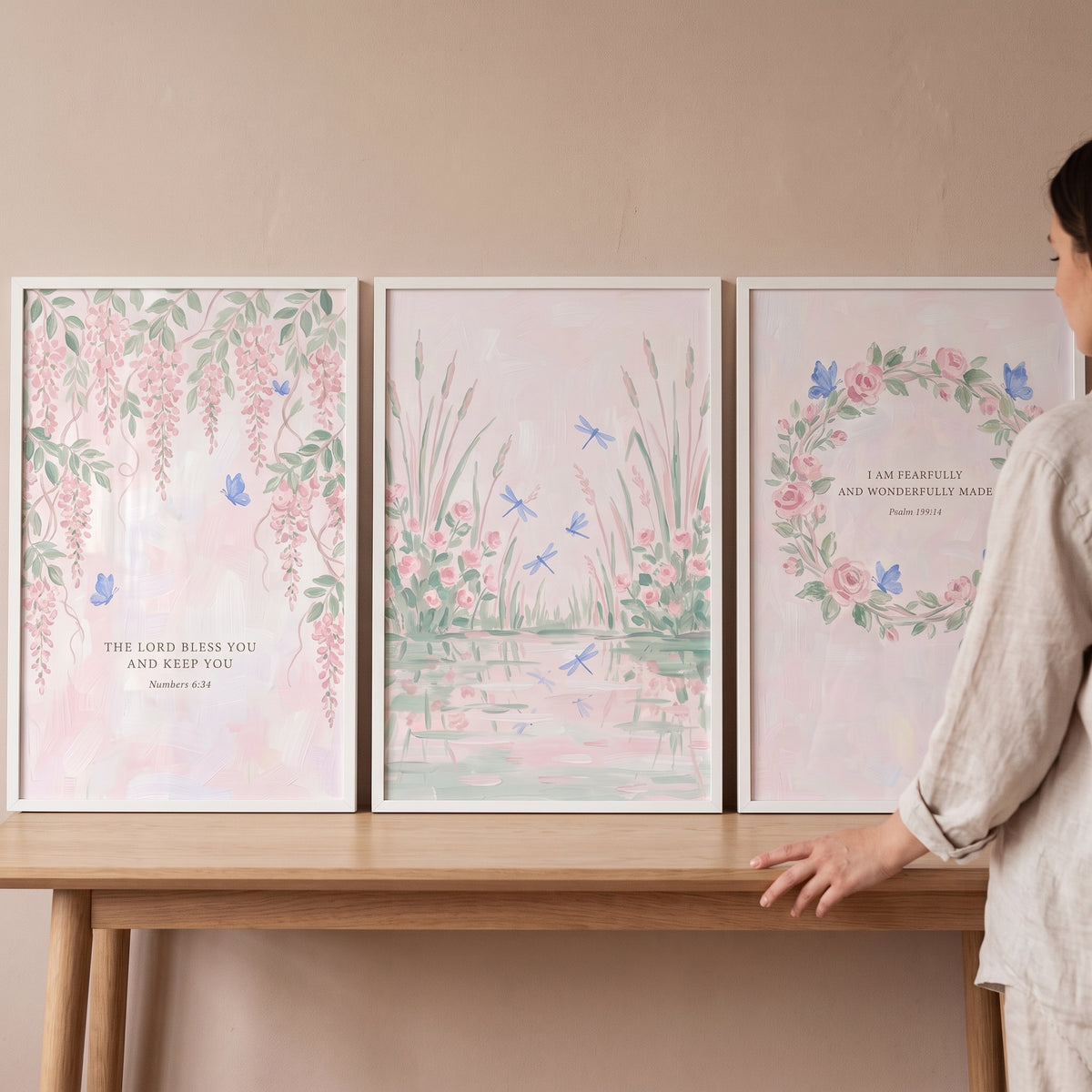 Dreamy blue winged insect wall art set of 3 framed pink floral prints with scripture on a wooden table next to a person.