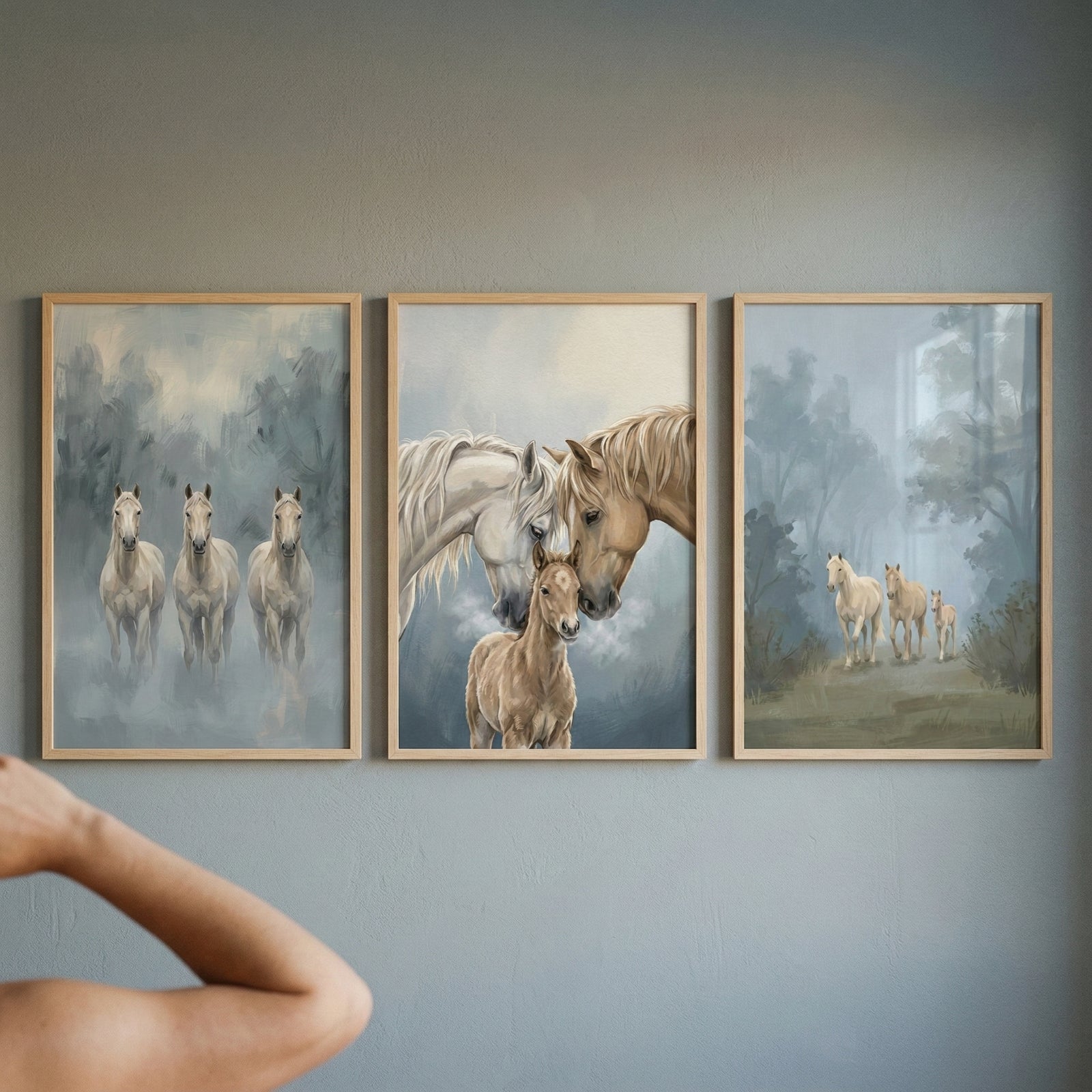 Three vertically-oriented, light-wood-framed artworks are hung in a row on a plain, grey wall. The pieces depict horses in a soft, ethereal style with a misty, atmospheric background. The leftmost artwork shows three pale horses standing together in a foggy woodland. The center piece is a close-up of two adult horses gently nuzzling a small foal. The rightmost artwork features three light-colored horses walking through a hazy landscape. An out-of-focus arm is visible in the foreground.