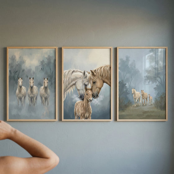 Three vertically-oriented, light-wood-framed artworks are hung in a row on a plain, grey wall. The pieces depict horses in a soft, ethereal style with a misty, atmospheric background. The leftmost artwork shows three pale horses standing together in a foggy woodland. The center piece is a close-up of two adult horses gently nuzzling a small foal. The rightmost artwork features three light-colored horses walking through a hazy landscape. An out-of-focus arm is visible in the foreground.