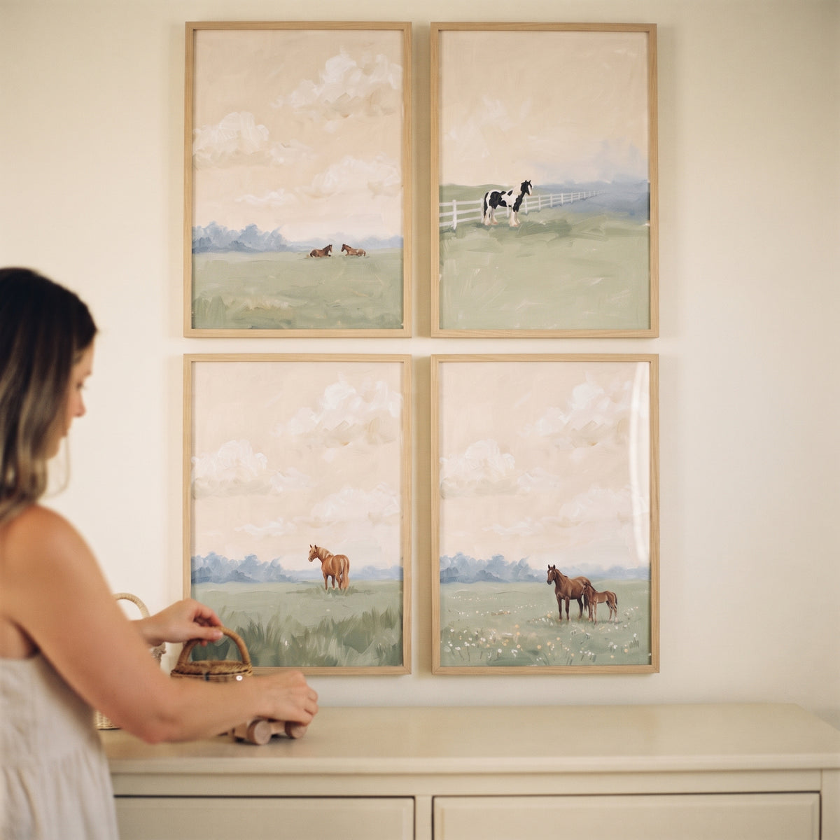 A woman stands by a dresser below a dreamy hazy field horses wall art set of 4 depicting horses in soft meadows.