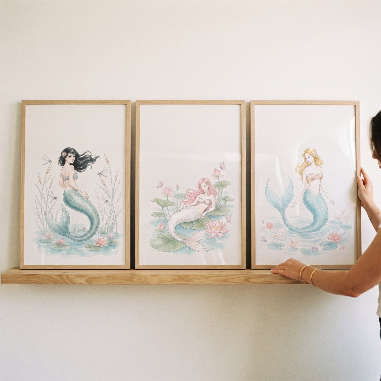 Hands adjust a dreamy pink lily mermaid wall art set of 3 displayed on a wooden shelf.