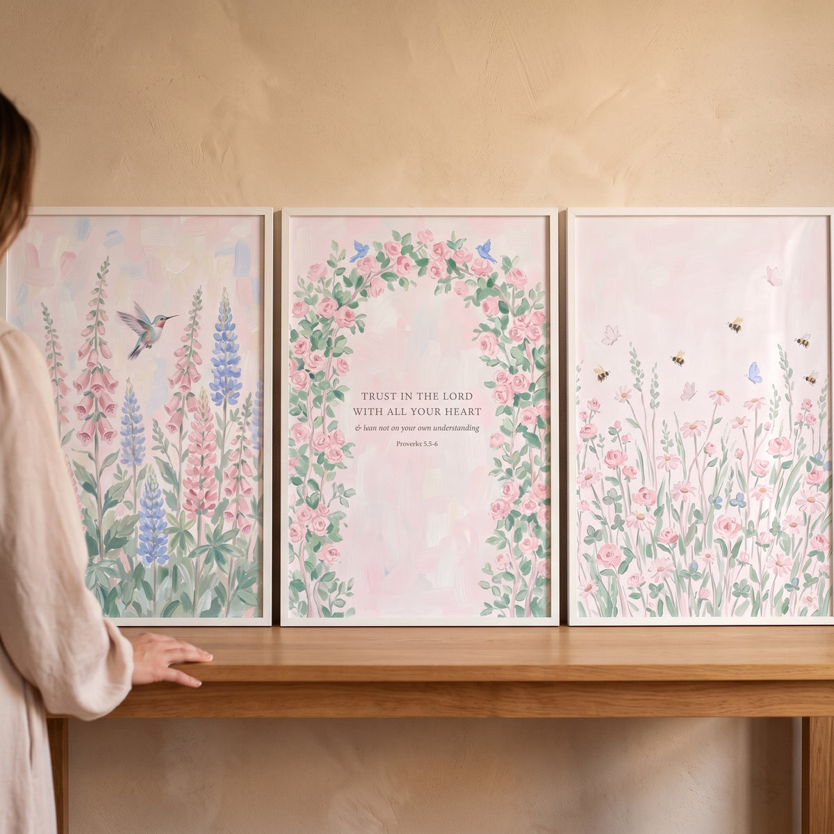 Dreamy watercolor pink garden wall art set of 3 with a hummingbird, floral arch, and religious verse on a wooden table.