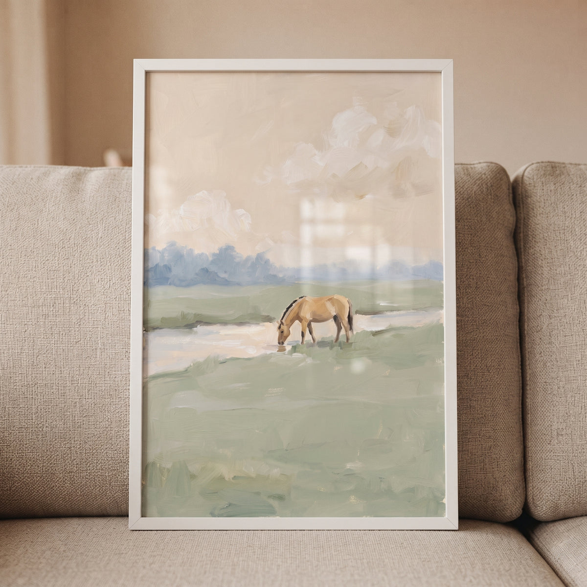 Drinking stream water horse wall art features a tan horse at a creek in a muted green landscape with a white frame.