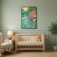 A serene nursery features a muted sage green wall adorned with a vertical framed artwork in the center. The artwork captures a vibrant jungle scene with a tiger leaning down to drink from a clear pool, its reflection rippling on the water's surface. The scene is lush with green foliage and delicate pink flowers under a bright yellow sun.
